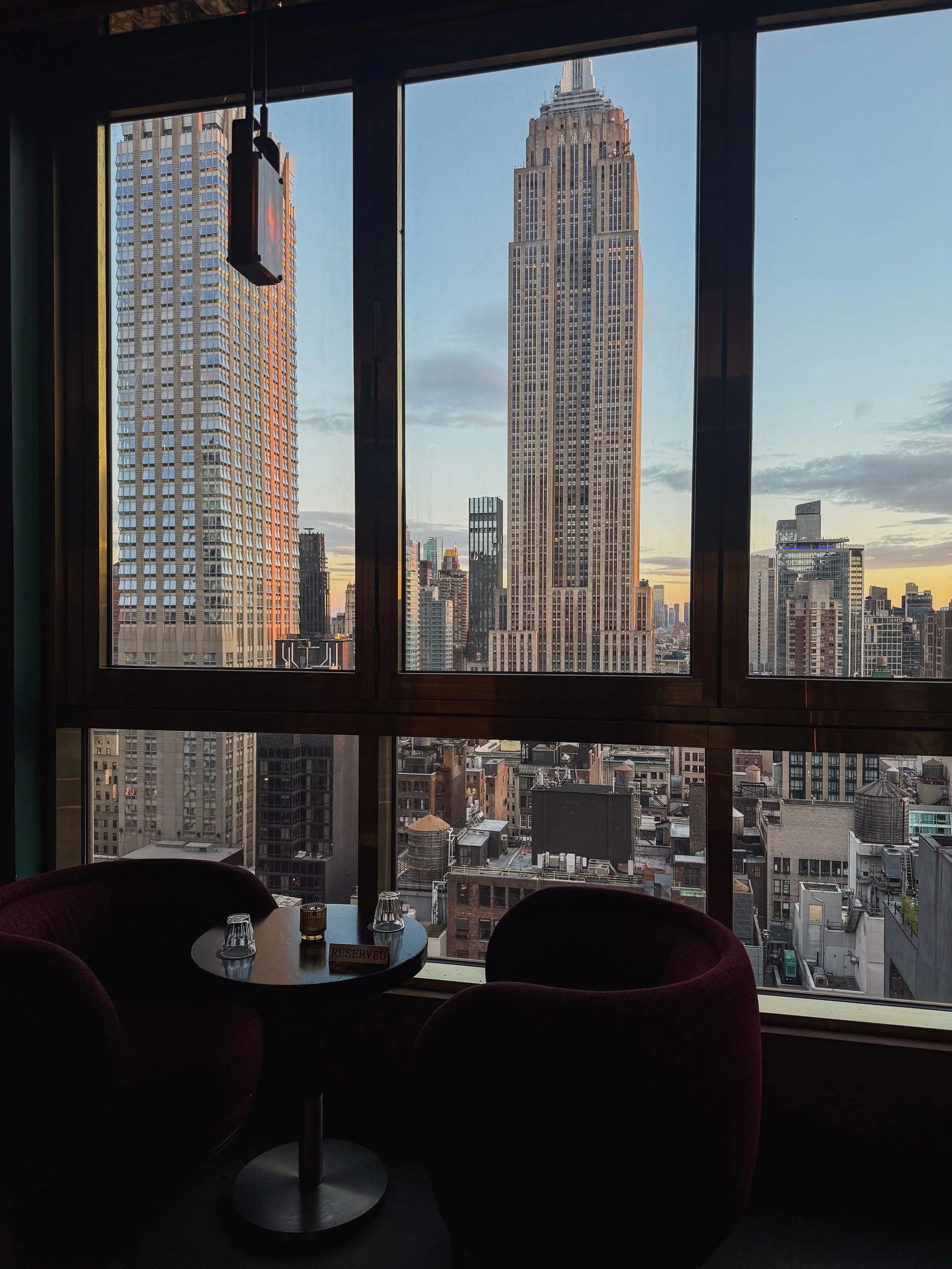 View of Manhattan's skyline from inside a high-rise building, with large windows showing the Empire State Building and other skyscrapers during sunset.