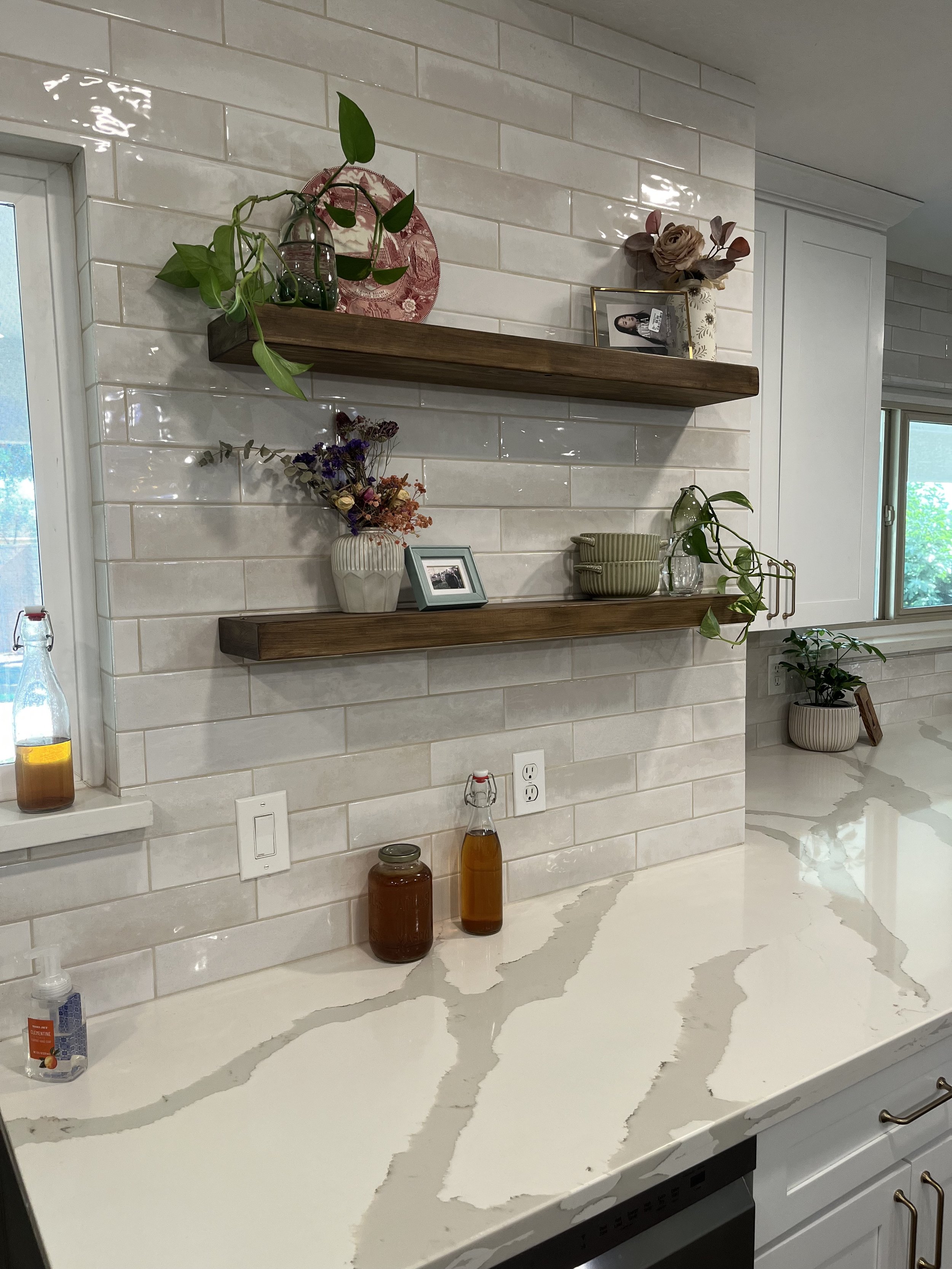A kitchen with a white marble countertop featuring grey streaks, a tiled white backsplash, and floating wooden shelves decorated with plants, picture frames, a flower vase, and jars. There are electrical outlets and a window on the left and right sides.