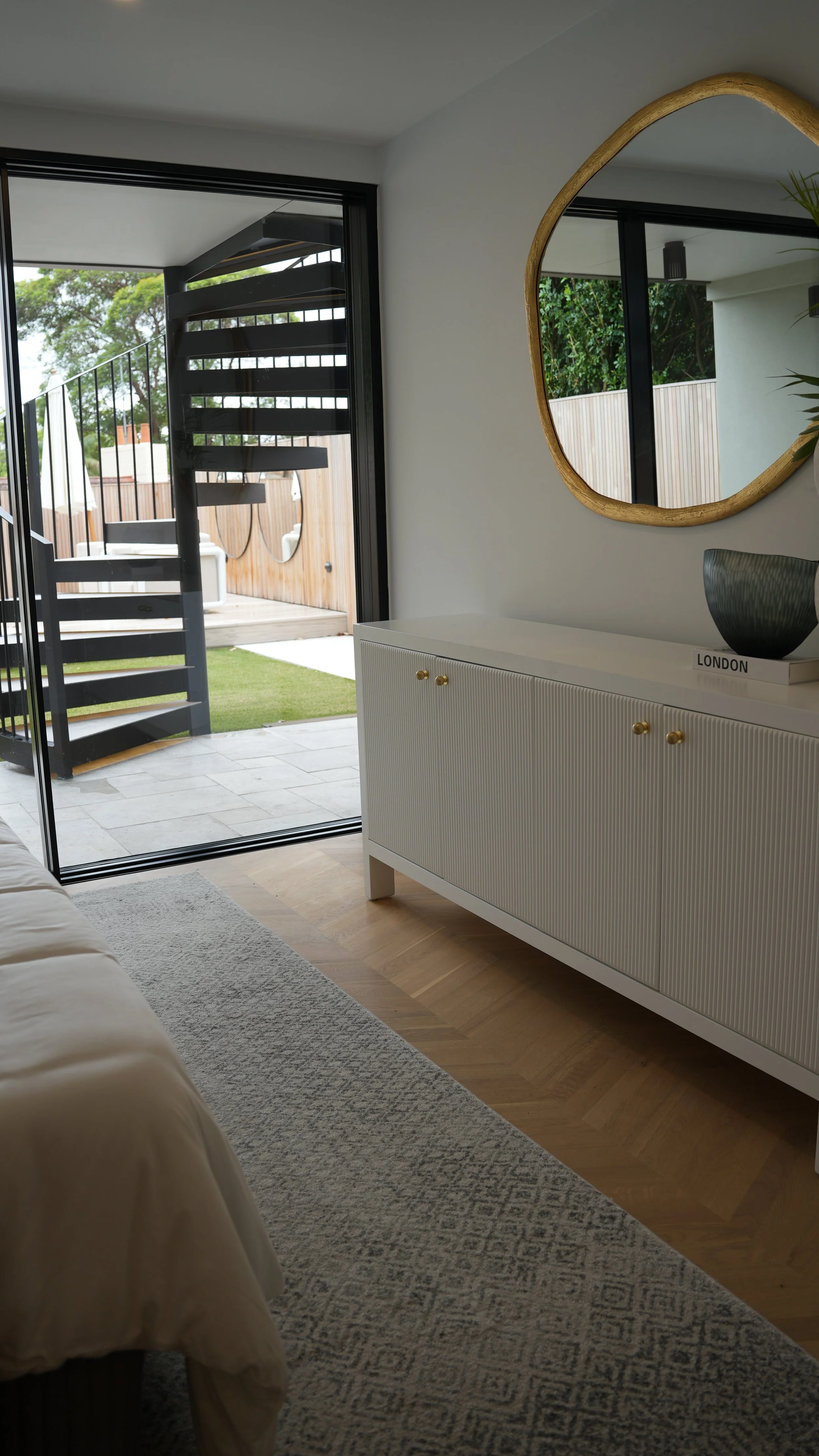 Interior view of a modern room with a white cabinet, a round mirror with a gold frame, and sliding glass doors leading to an outdoor patio with a spiral staircase, wooden fencing, and greenery.