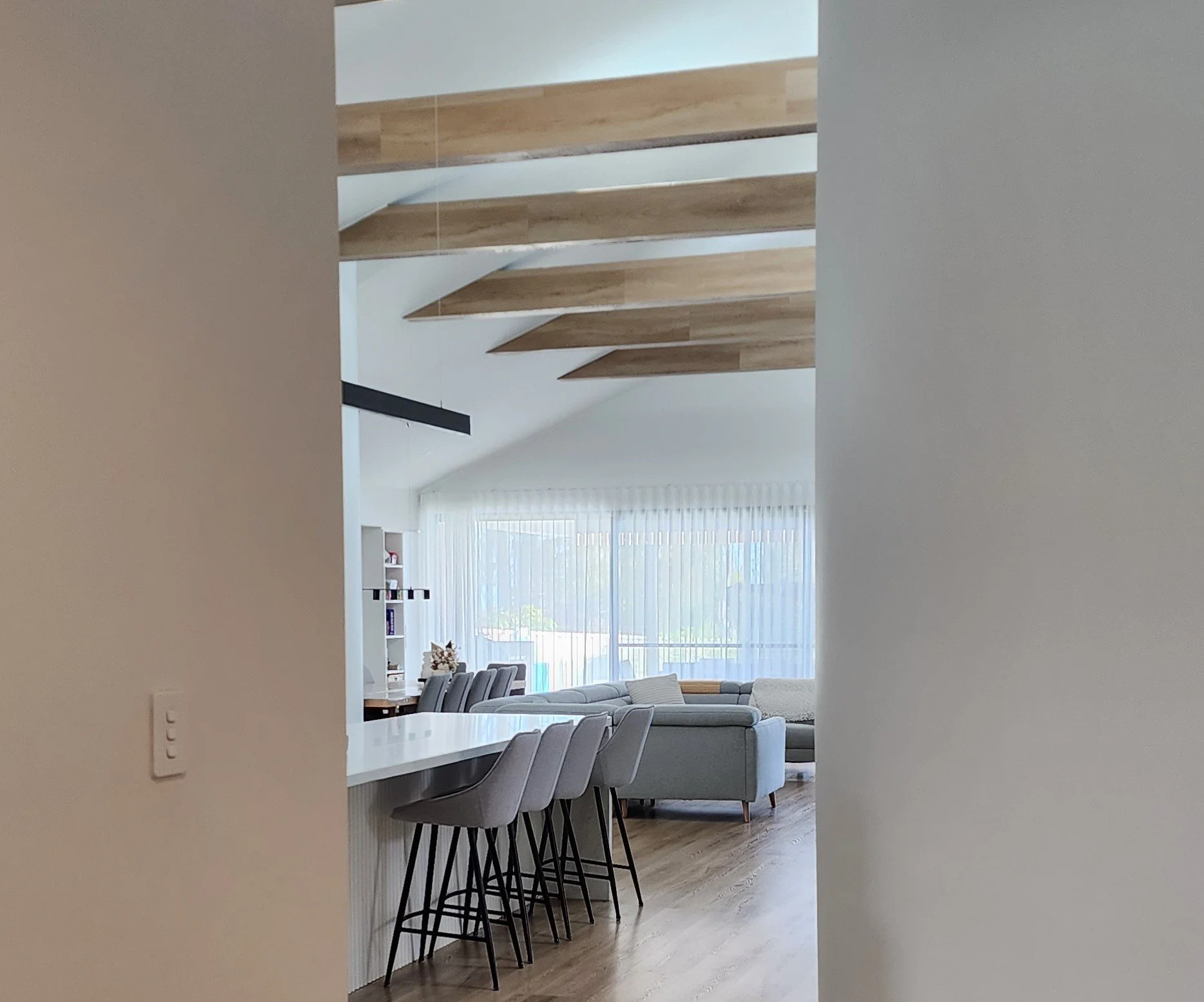 Open-concept living room and kitchen area with a breakfast bar and bar stools, featuring a modern interior with large windows and sunlight.