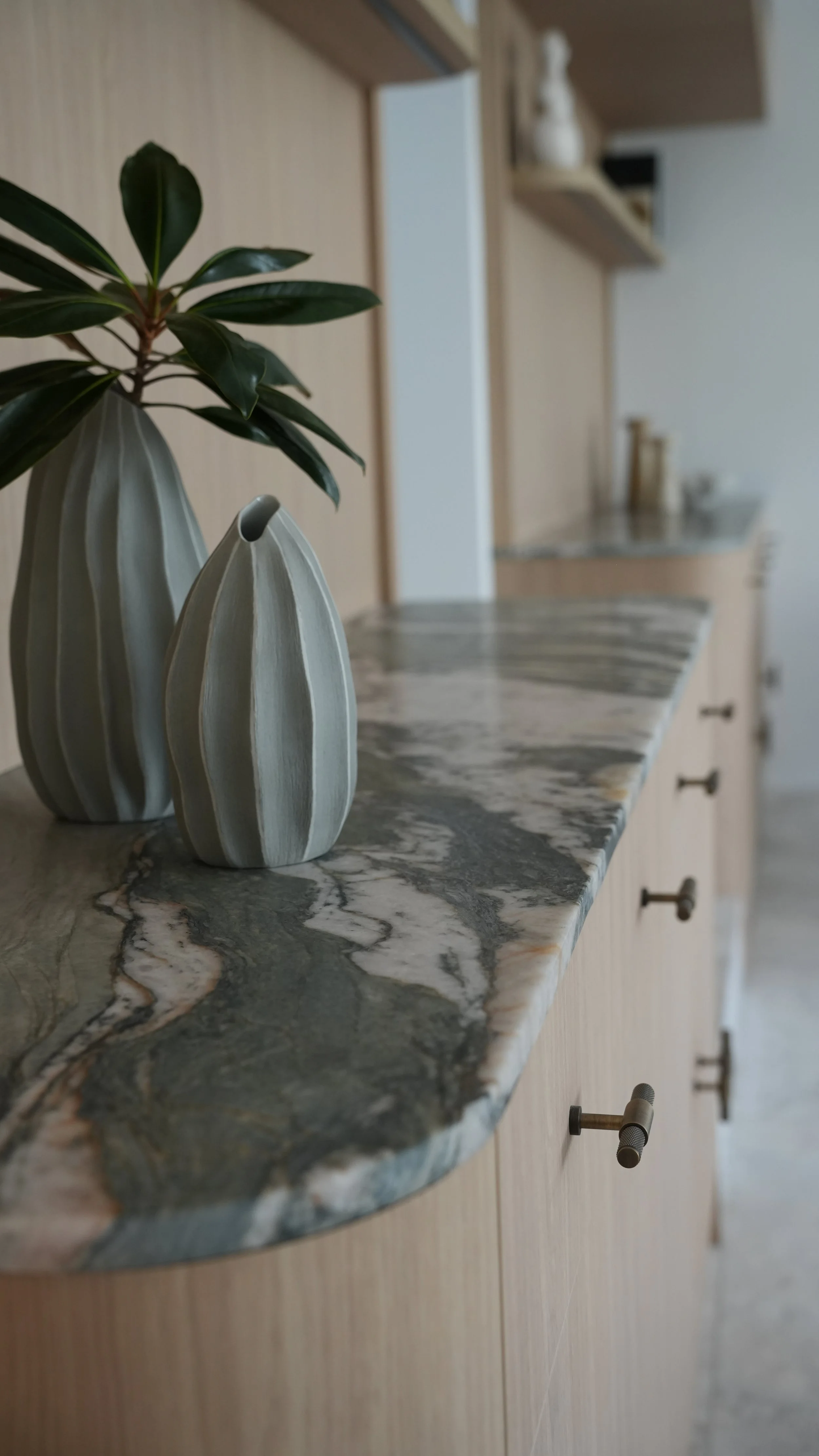 Close-up of a marble kitchen countertop with two decorative ceramic vases and a houseplant. In the background, kitchen shelves with vases and a countertop with jars and utensils are visible.