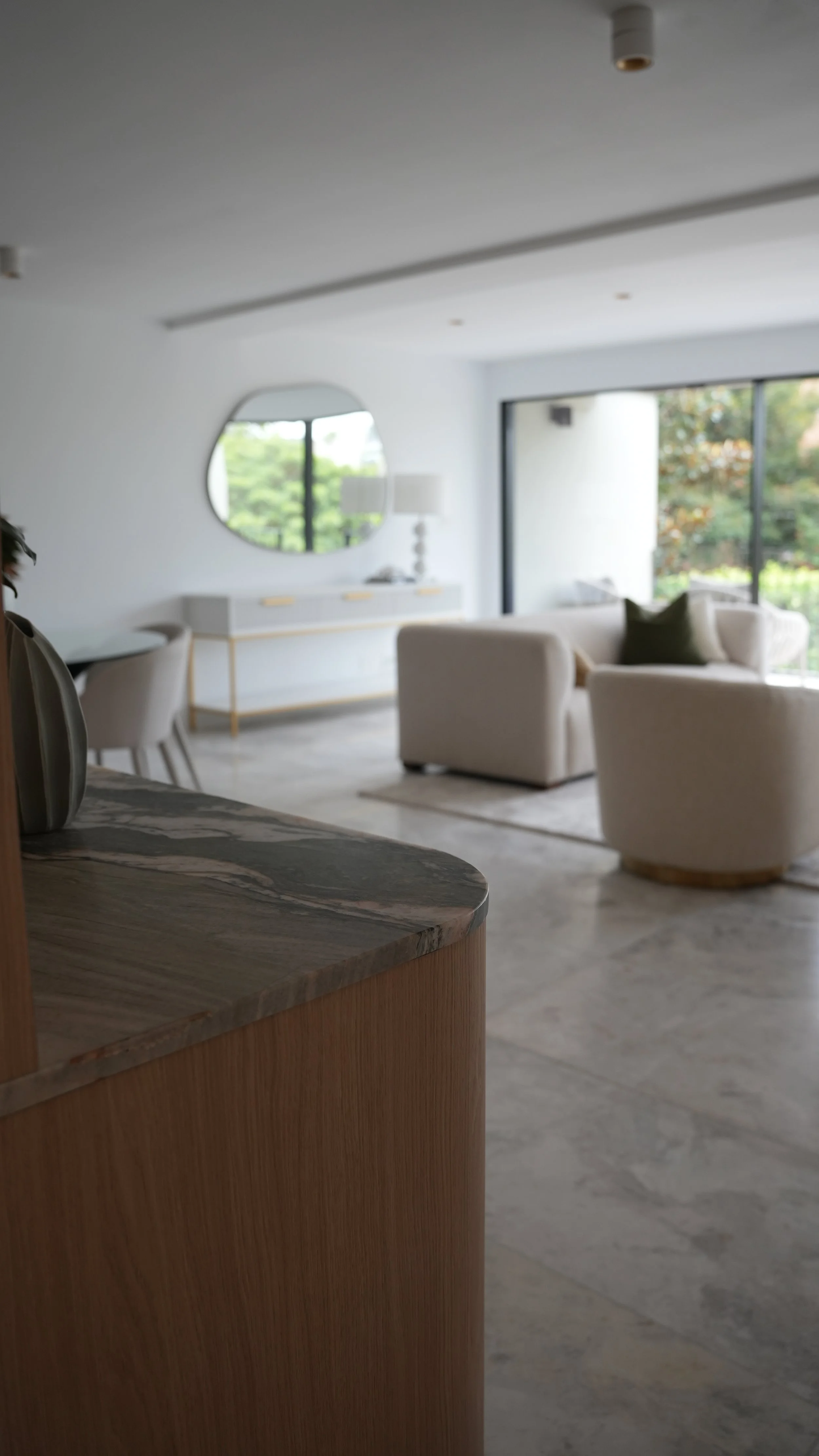 A modern living room with white sofas, a sideboard with a mirror, large window with greenery outside, and a marble countertop in the foreground.