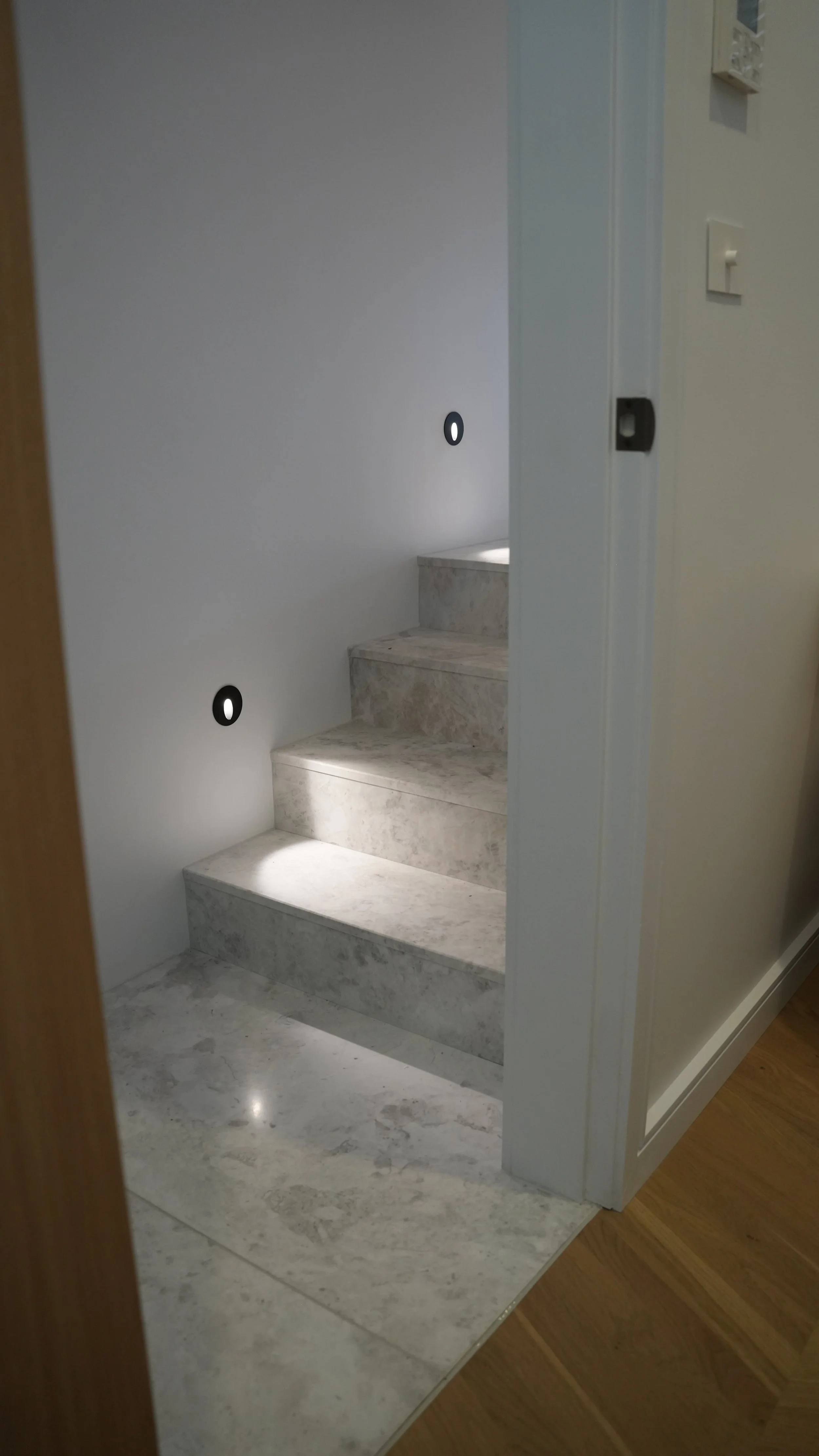 A marble staircase with three steps, installed in an indoor space with white walls, grayish marble flooring, and small black circular wall lights on the wall beside the stairs.