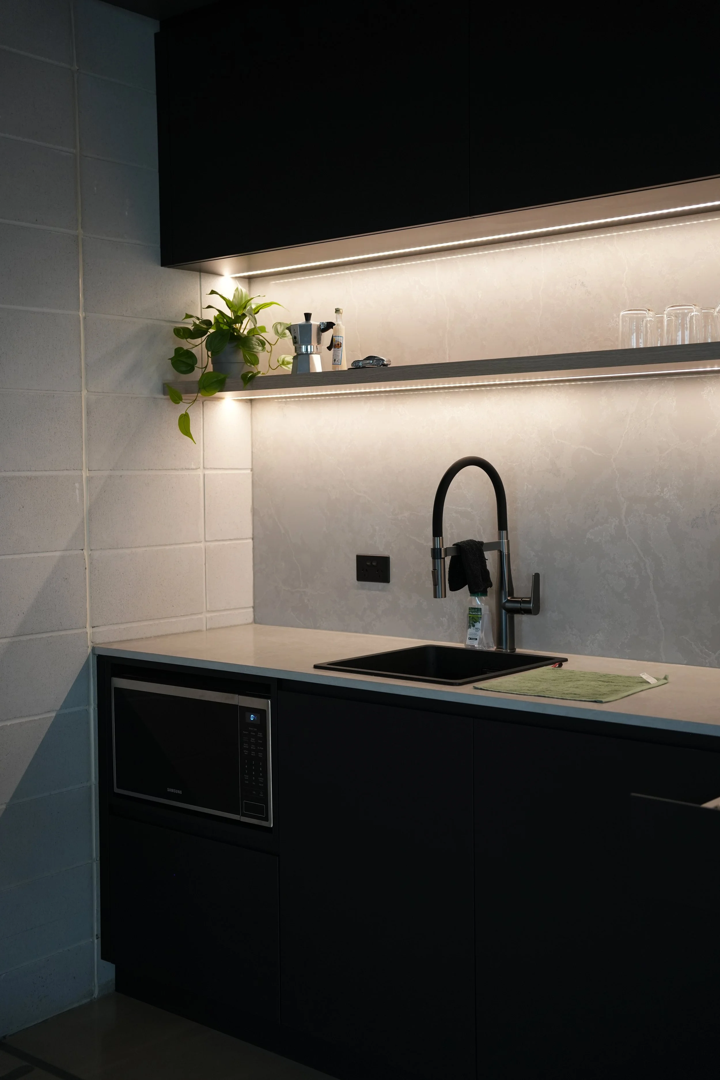 Modern kitchen with black cabinetry, a gray countertop, and a sink with a black faucet. Open shelves above the sink hold a potted plant, a moka pot, a small bottle, a toy car, and glassware. Under the counter, there's a microwave oven.