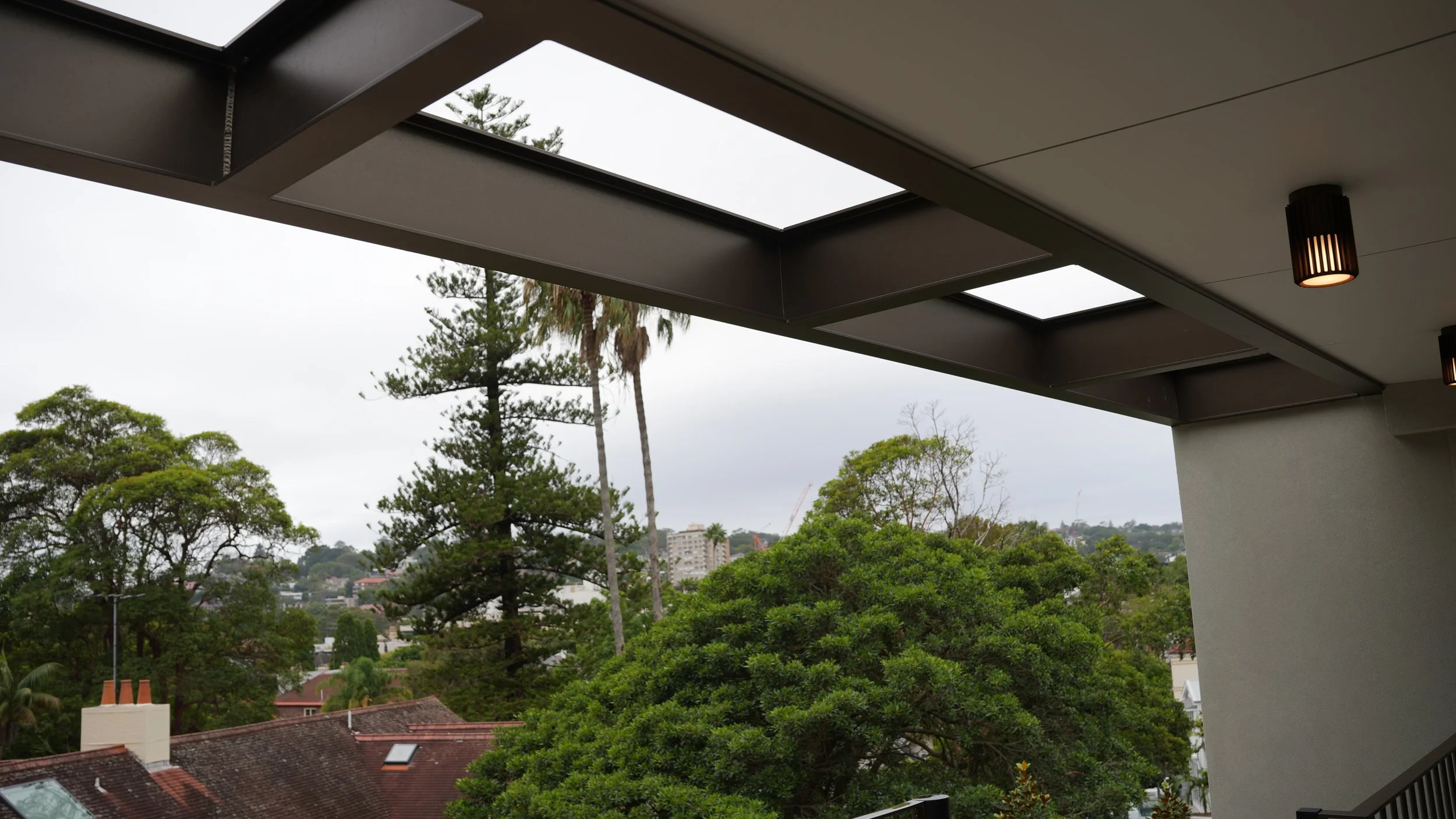 Overcast sky viewed through ceiling skylights on a balcony, with green trees and rooftops in the distance.