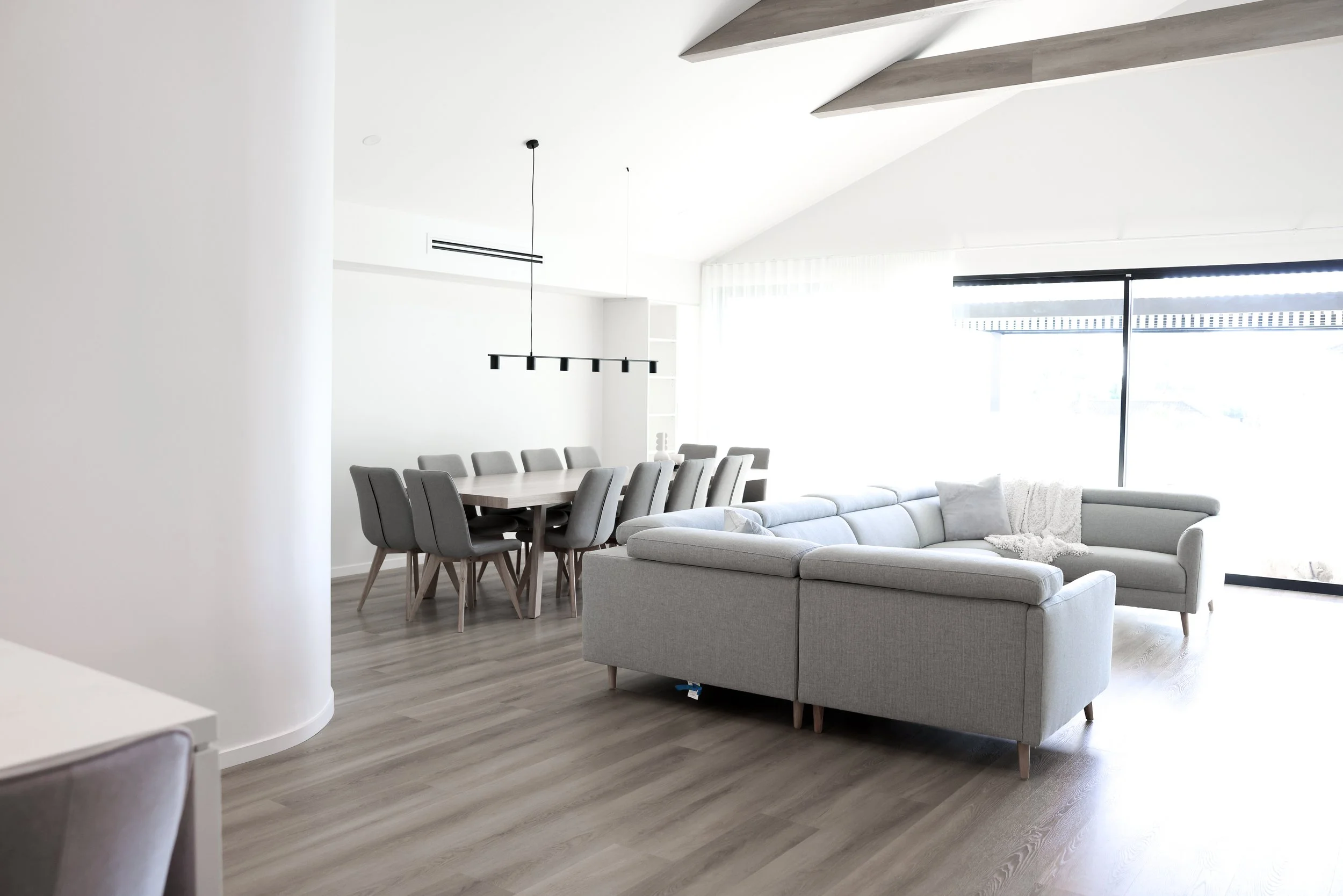 Modern, bright living room with a large sectional sofa, a dining table with chairs, and a minimalist chandelier, illuminated by large windows with sheer curtains.