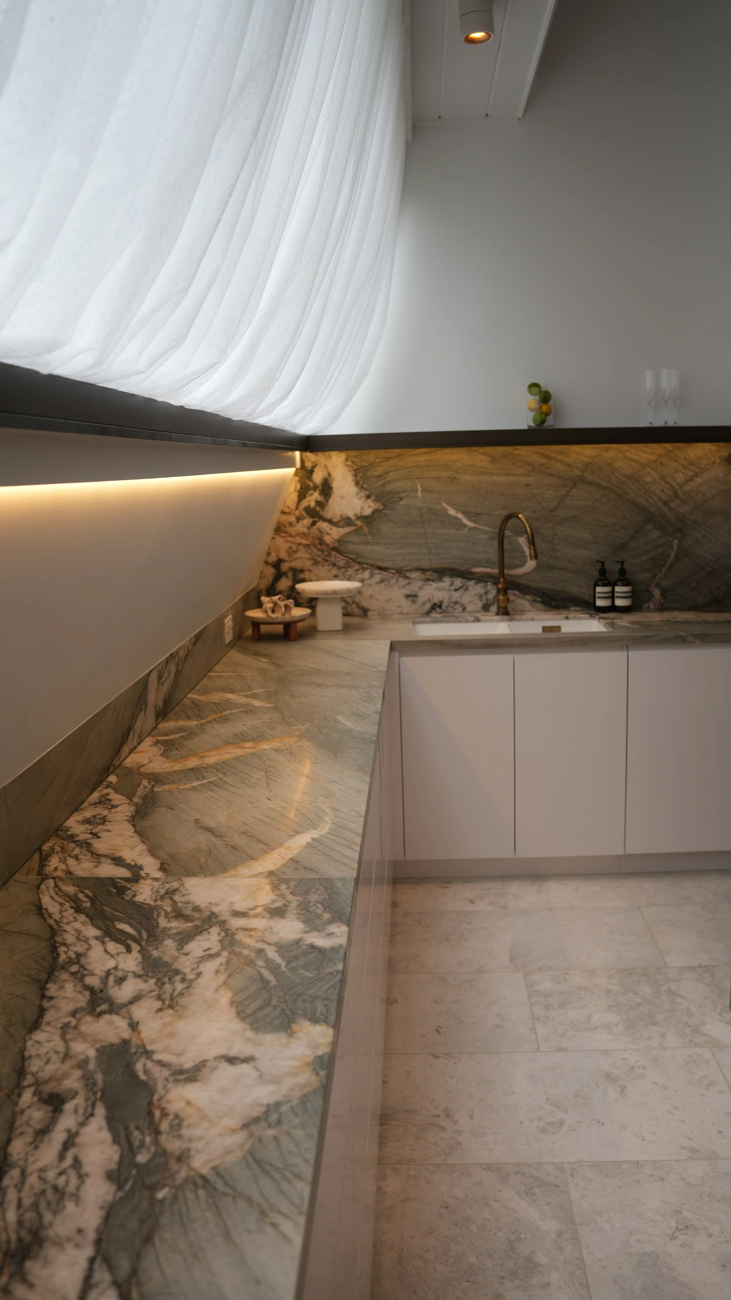 A modern kitchen with a marble countertop, a sink with a bronze faucet, white cabinets, and decorative items including two glasses, a small terrarium, and soap dispensers. Sheer curtains cover a large window, and the space has recessed lighting.