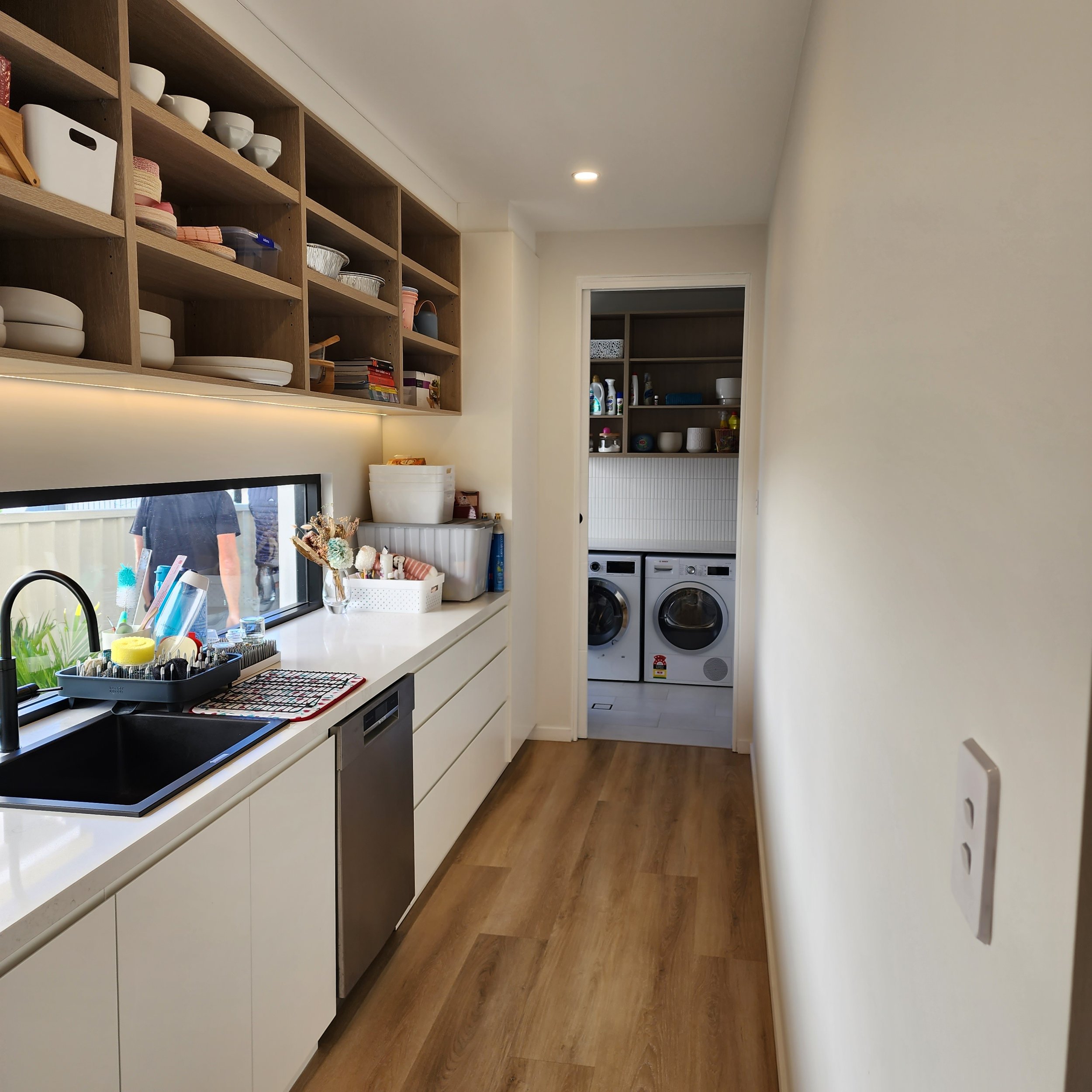 A modern laundry room with white cabinets, a long white countertop, and wooden flooring. Cabinets above hold dishes and kitchenware, while open shelves contain cleaning supplies and other household items. A window above the sink area provides natural