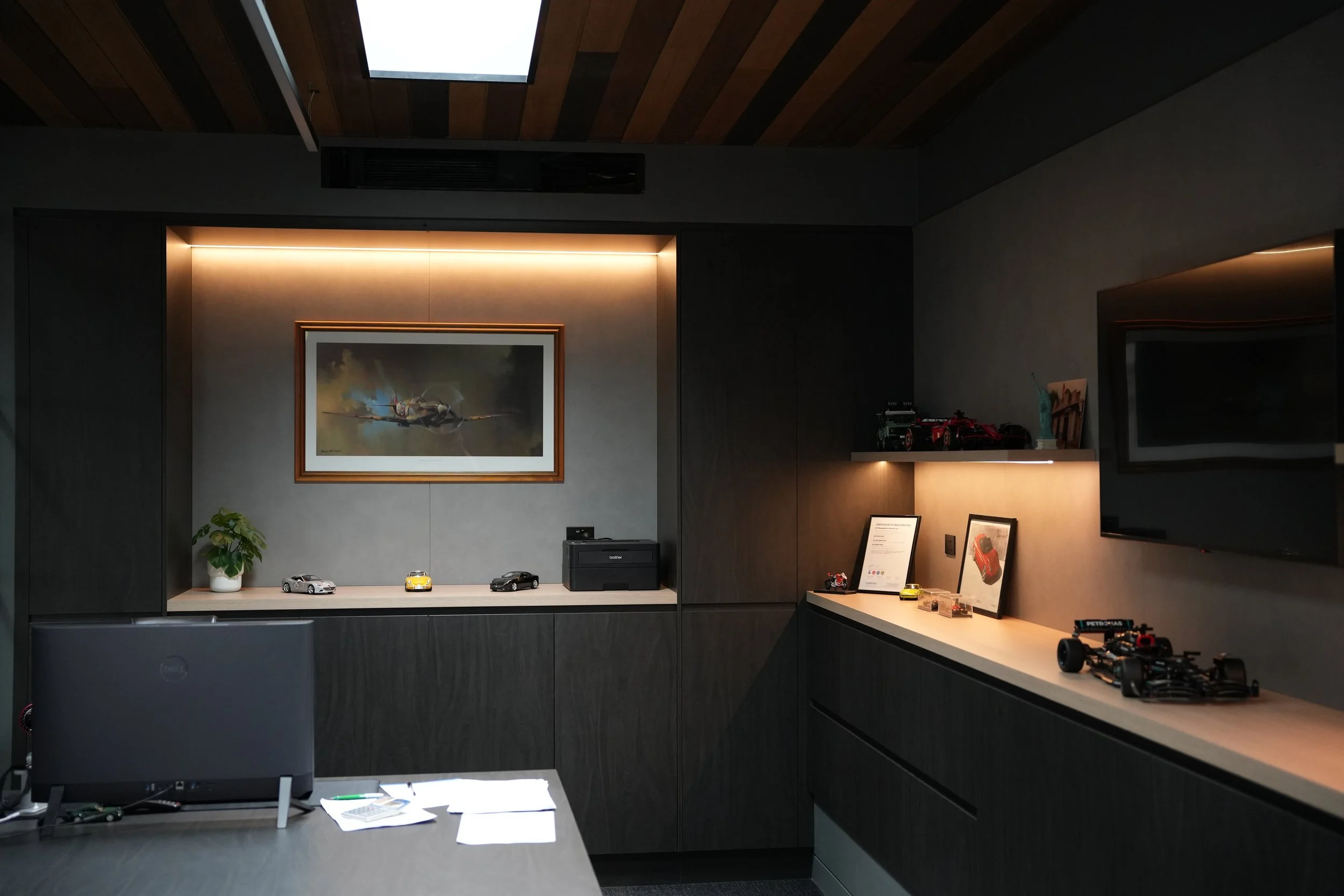 A modern office with a wooden ceiling and dark walls. On the wall, there is a framed picture of a fighter jet airplane. Below the picture, a wooden shelf holds several small car models, a potted plant, and some framed photographs or artwork. To the right, a section of a countertop displays a framed document and more car models. The lighting is warm and accentuates the items on display.