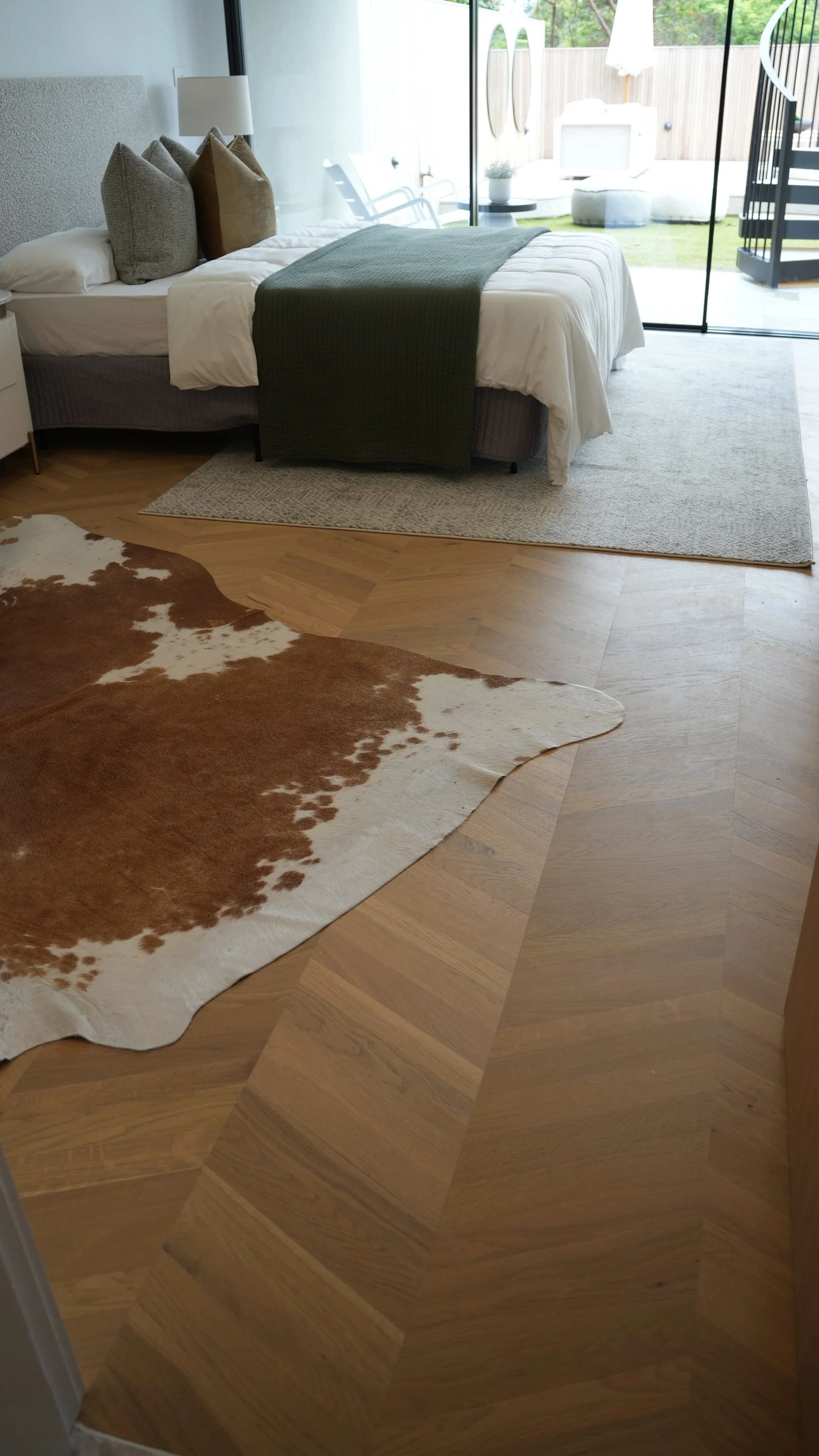 A bedroom with a bed, pillows, and a view of an outdoor patio through glass sliding doors. The floor is wooden with a cowhide rug partially visible.