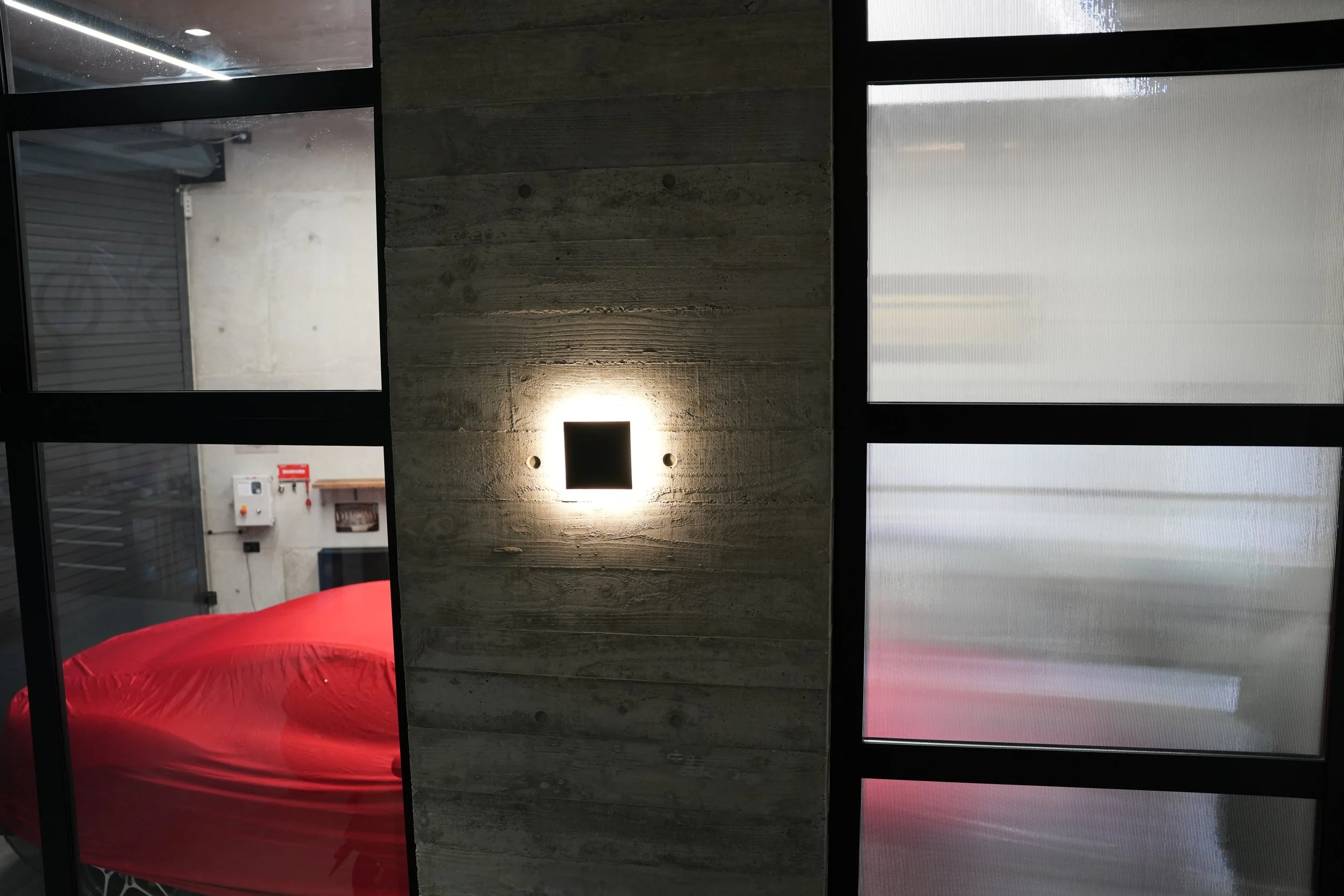Interior view of a modern building with large windows and a textured concrete wall illuminated by a square wall light. There is a red fabric-covered surface or chair and some utility controls on the wall. The view includes part of an outdoor garage o