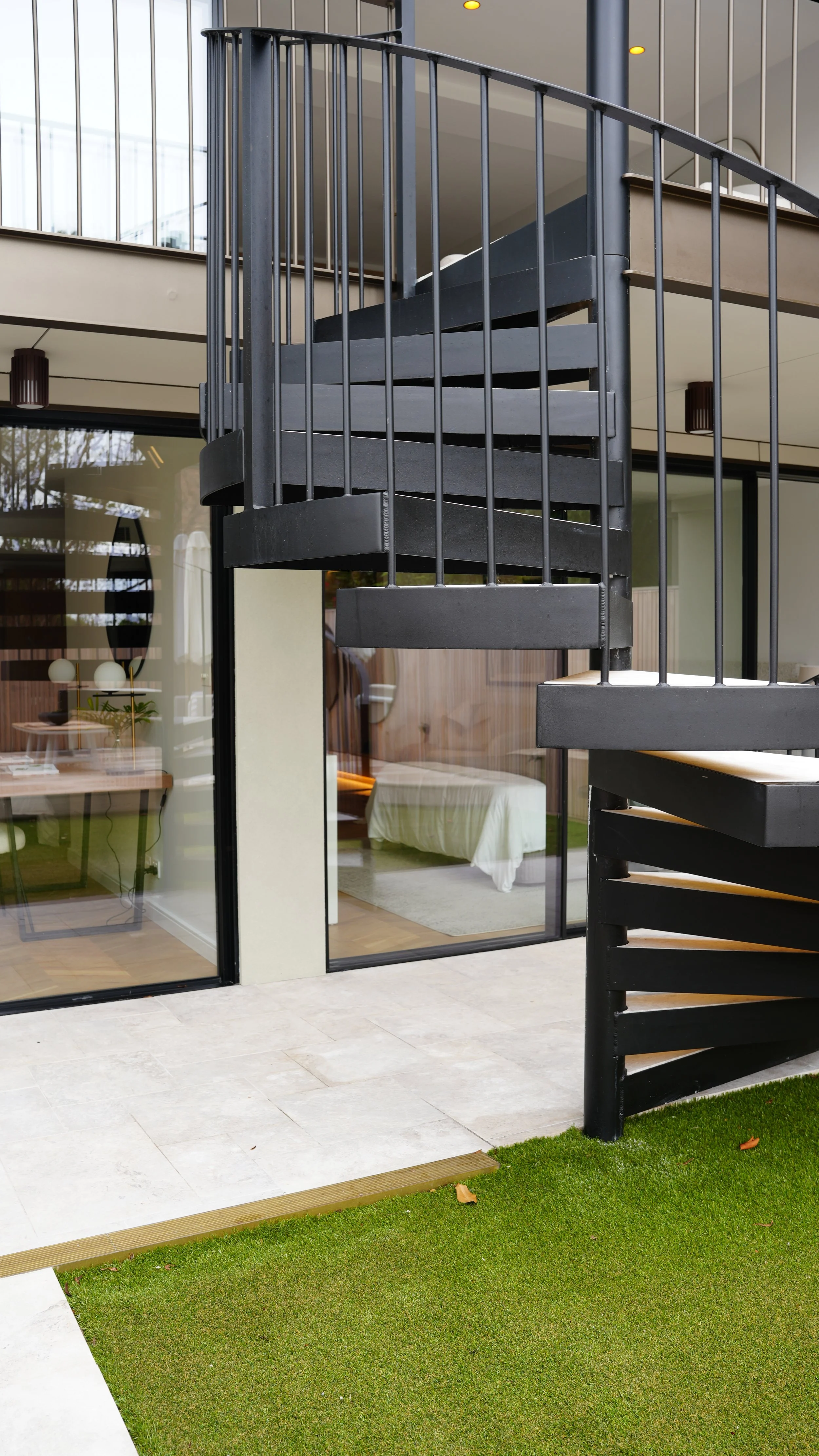 Photo of a modern black spiral staircase outside a house, with glass balcony railings, sliding glass doors leading to the interior bedroom, and a small grass lawn.