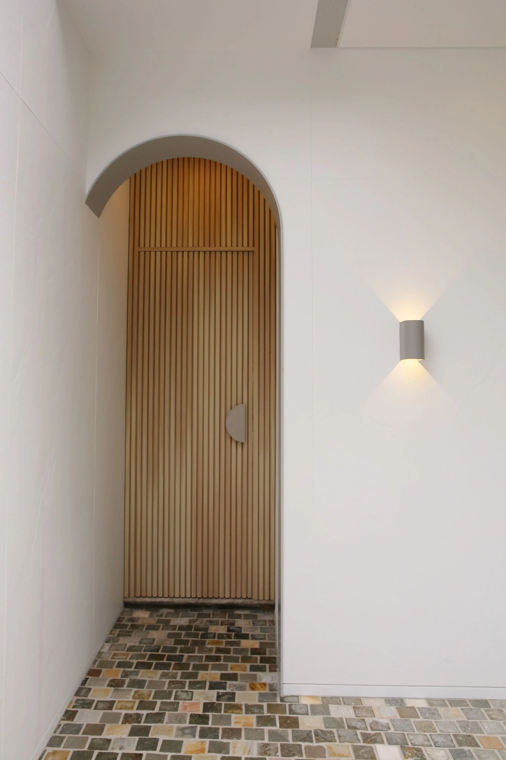 A doorway with a curved top and wooden slats, set in a white wall. To the right, a modern wall-mounted light fixture is turned on, casting light upward and downward. The floor is made of multicolored rectangular tiles.