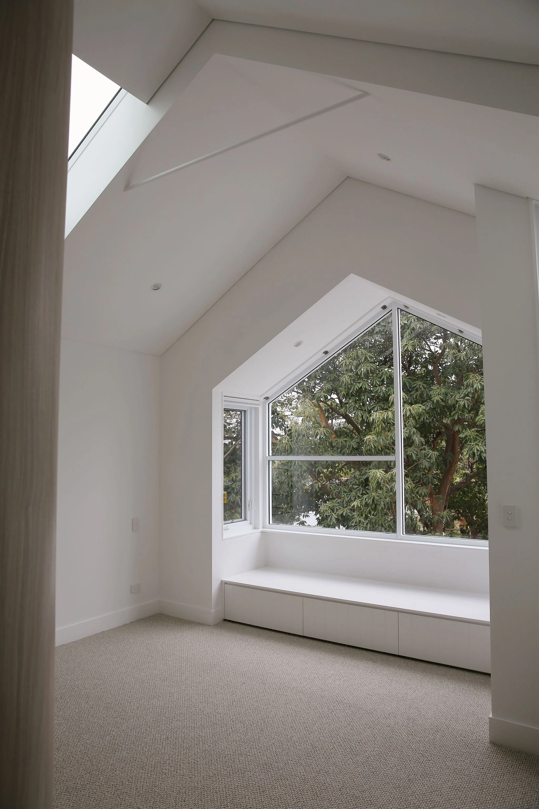 Interior room with large window, white walls, beige carpet, built-in window seat, and a partial view of trees outside.