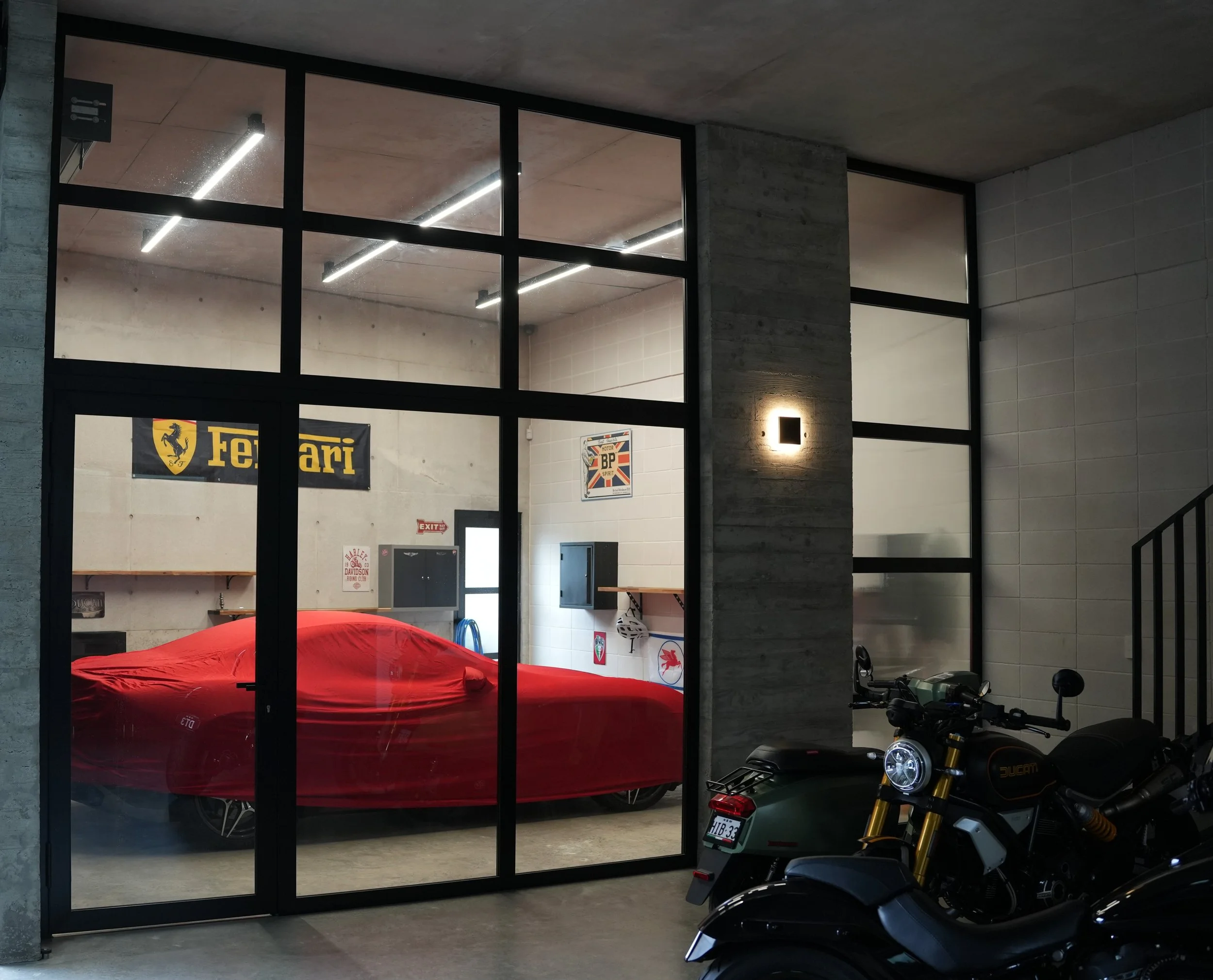 View of a modern indoor space with large glass windows showing a red vehicle covered with a protective cover inside. The room features a concrete wall with Ferrari banners, a motorcycle parked in the foreground, and industrial-style lighting on the c