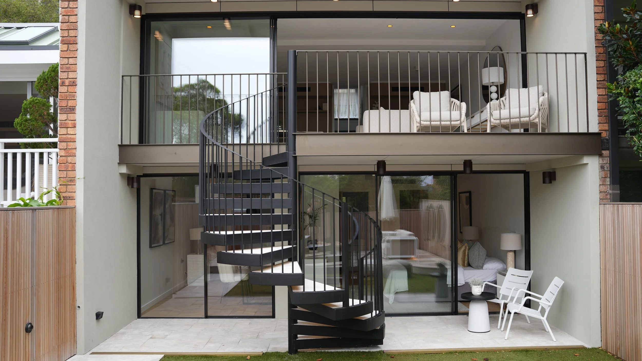 Exterior view of a modern two-story house with a glass door and balcony. The balcony has light-colored chairs and a lamp, and a spiral staircase leads to the upper floor. The ground level features a seating area inside visible through large glass doo