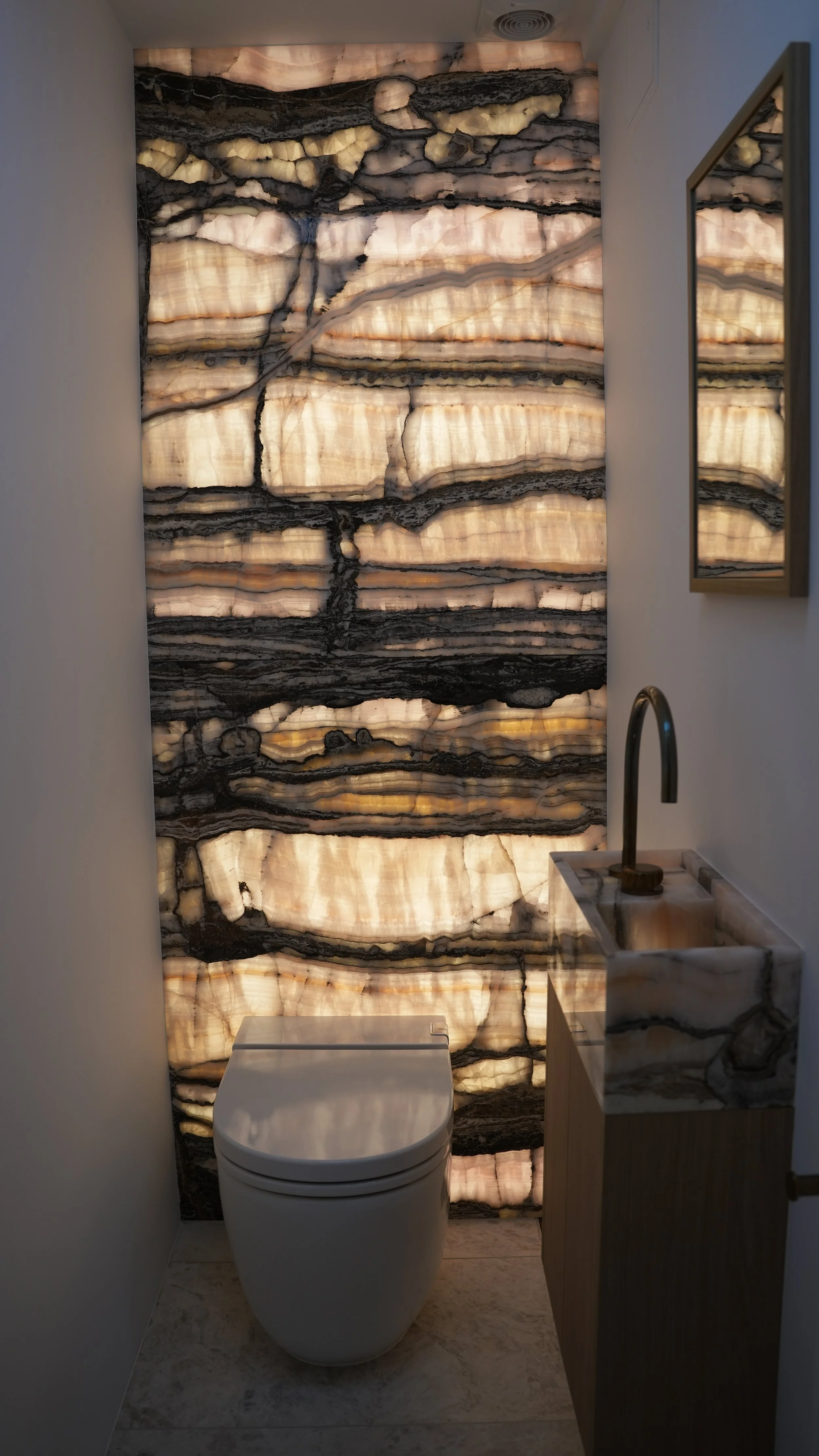 A modern bathroom with backlit onyx stone wall, a white toilet, a marble sink with a gold faucet, a mirror, and a marble countertop.