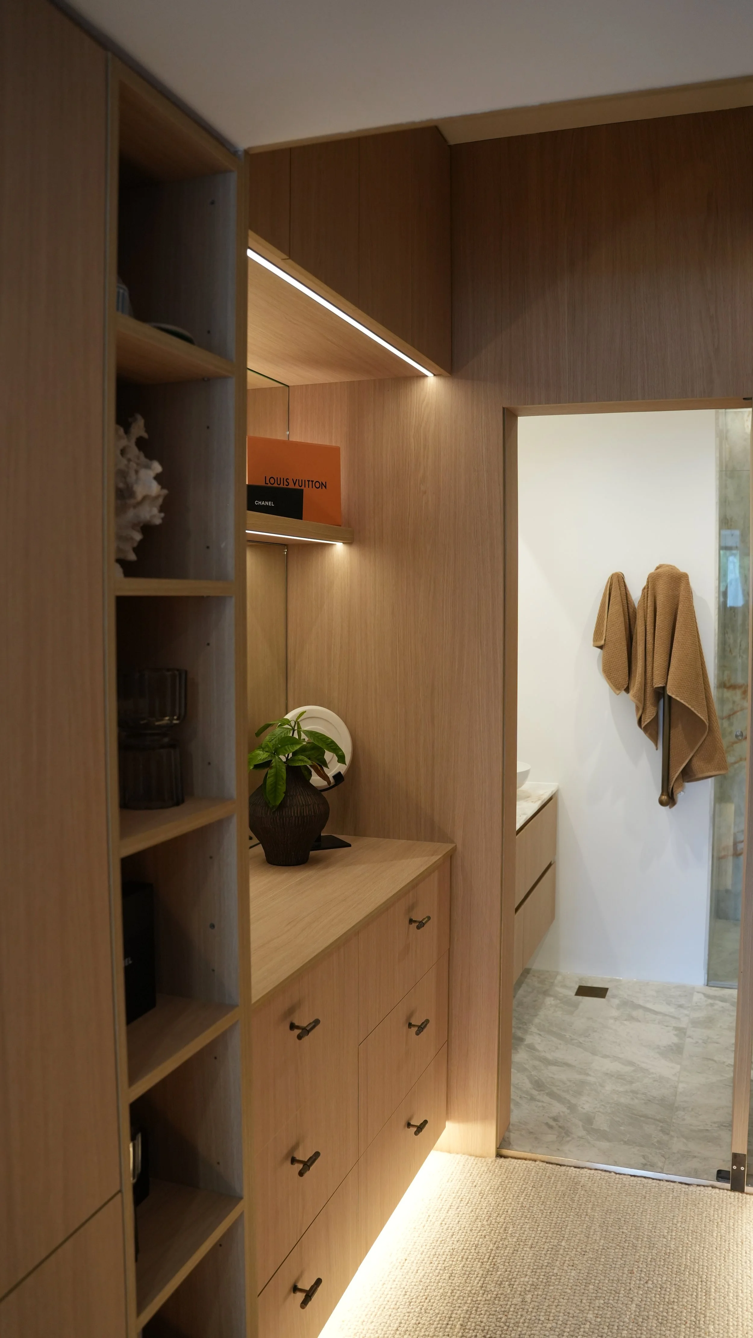 A wooden storage cabinet and shelves in a bathroom or dressing area, with a plant and decorative items on top, and towels hanging on the wall.