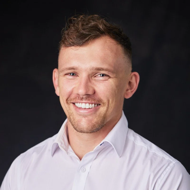 A smiling man with short, light brown hair, blue eyes, wearing a white button-up shirt, against a dark background.