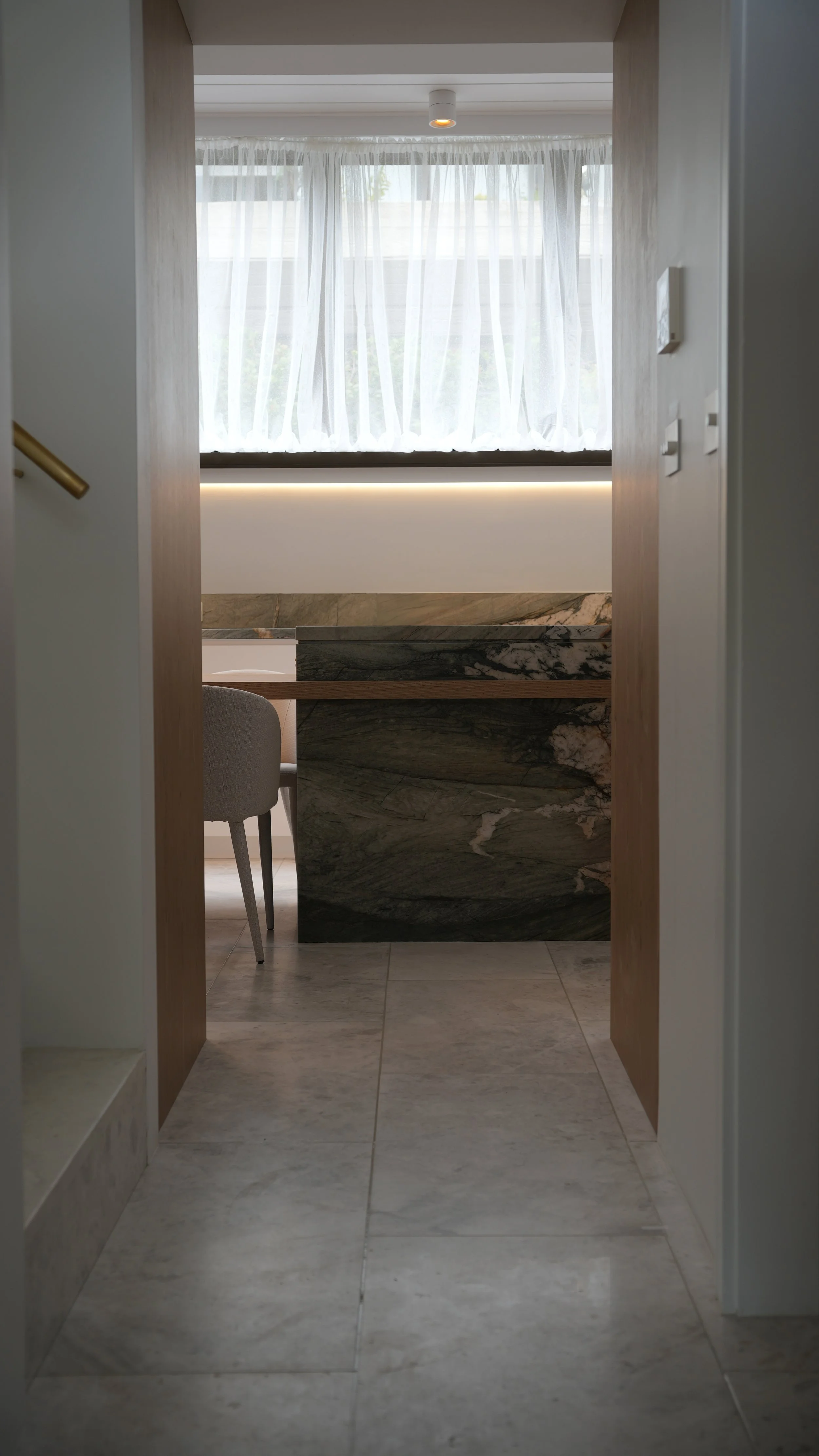 View through a doorway into a room with a big window and white curtains, a marble kitchen island, and a chair.