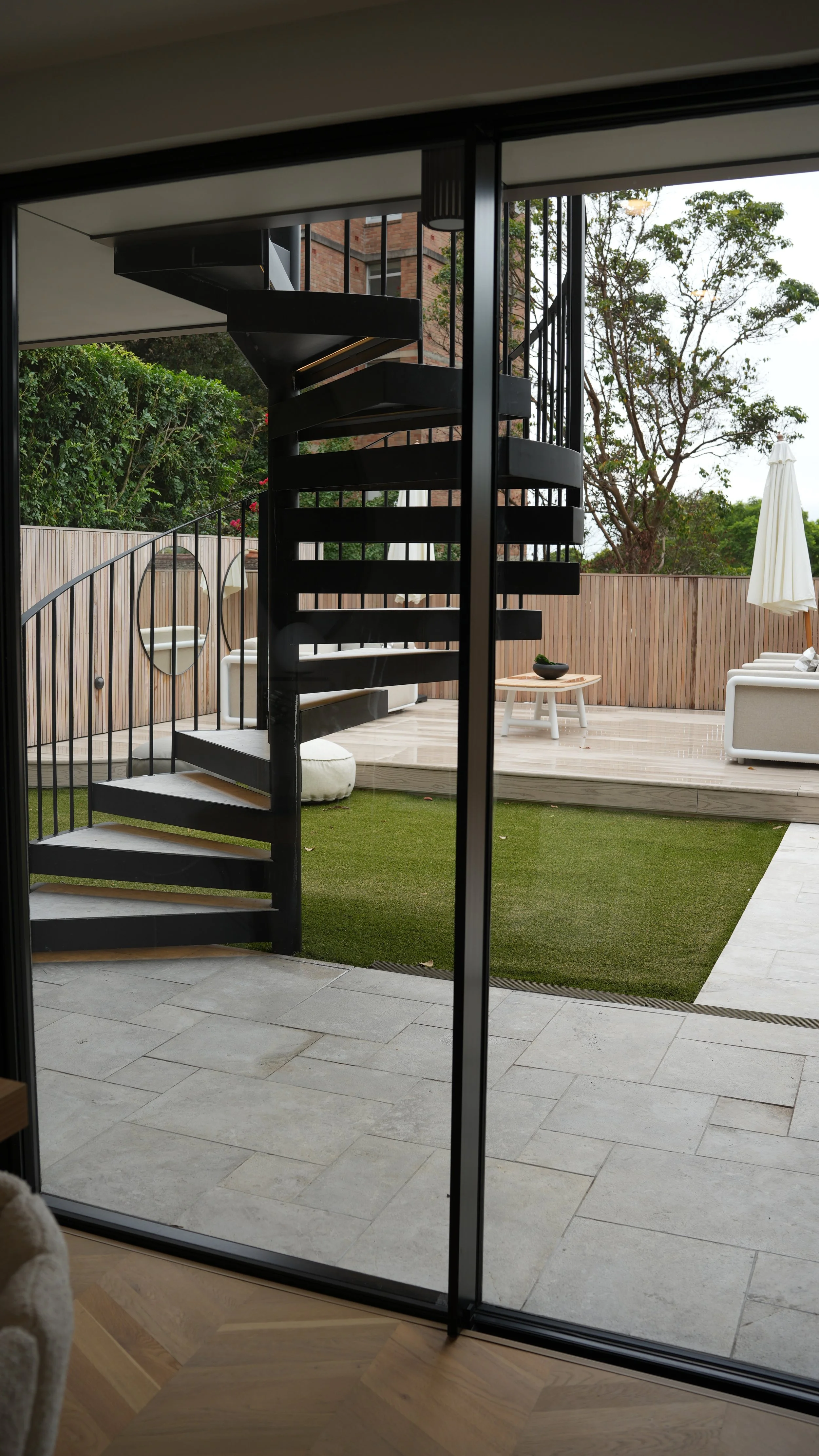 View of an outdoor patio area with modern furniture, a spiral staircase, and a wooden fence with trees in the background.