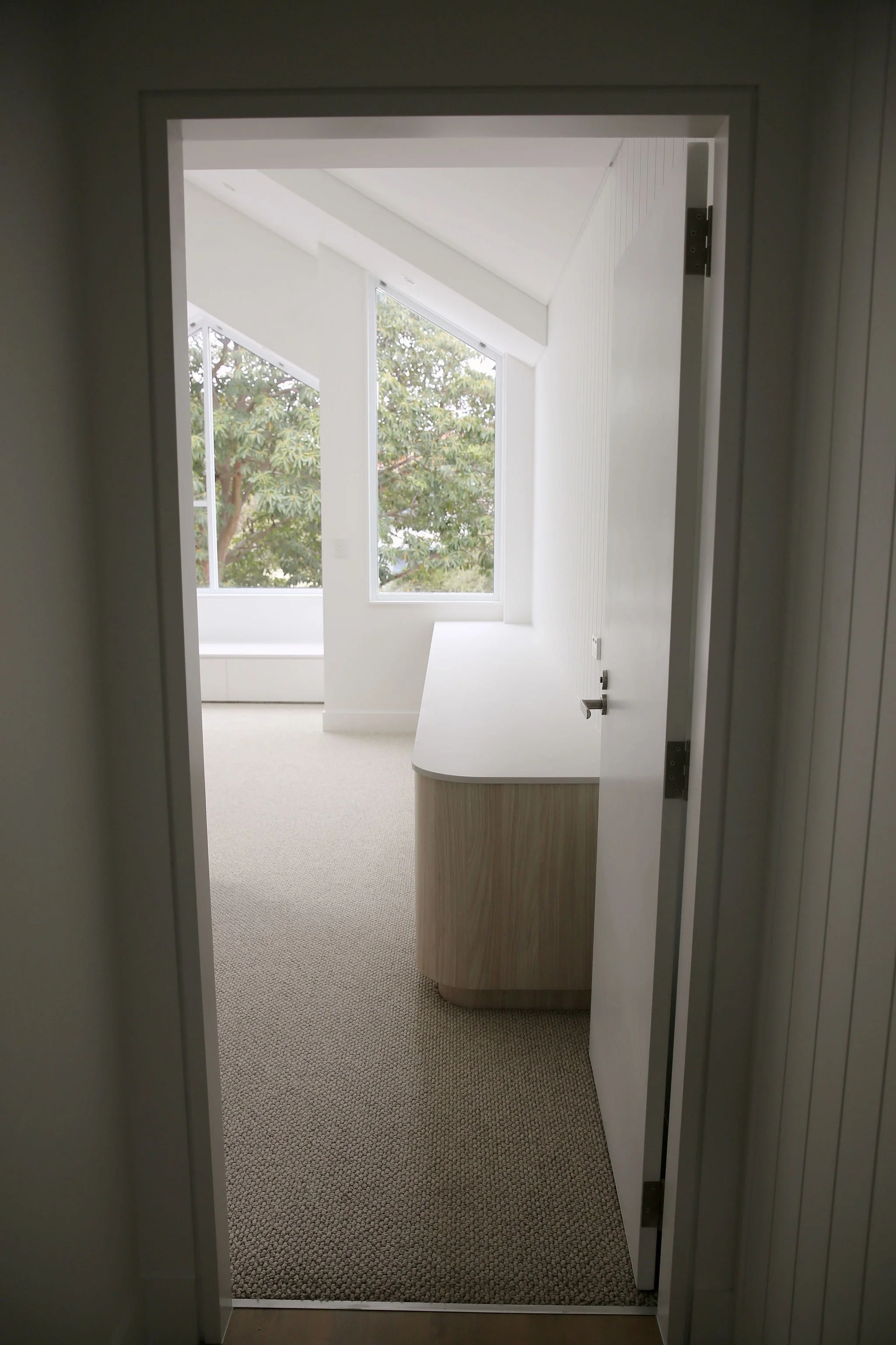 View through a doorway into a bright room with large windows and a sloped ceiling, featuring a curved wooden cabinet or desk.