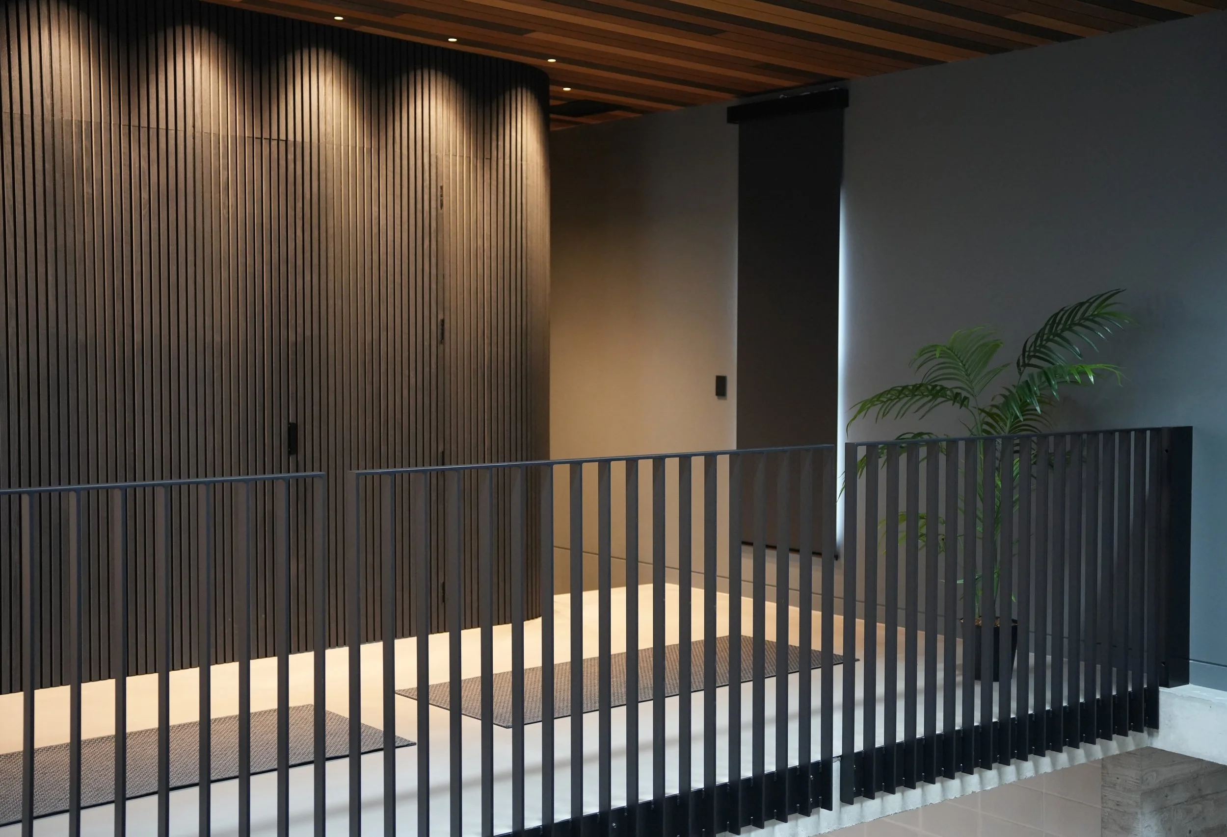 Modern interior space with a wooden slat wall, black railing, indoor plant, and ceiling lights.