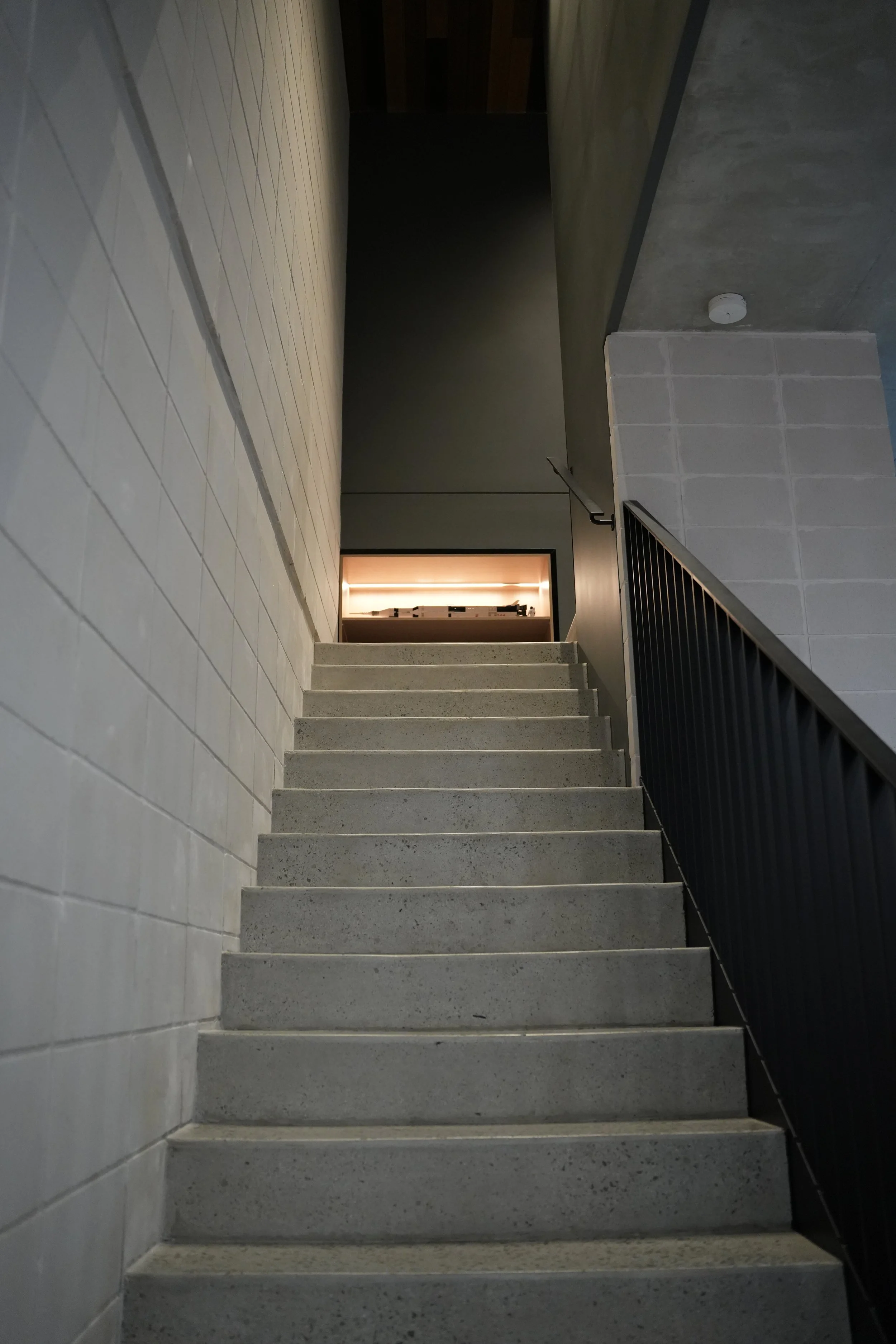 A staircase made of concrete with a black metal railing on the right side. The walls are decorated with light-colored tiles on the left, and plain gray concrete on the right. At the top of the staircase, there is a doorway with warm lighting visible 