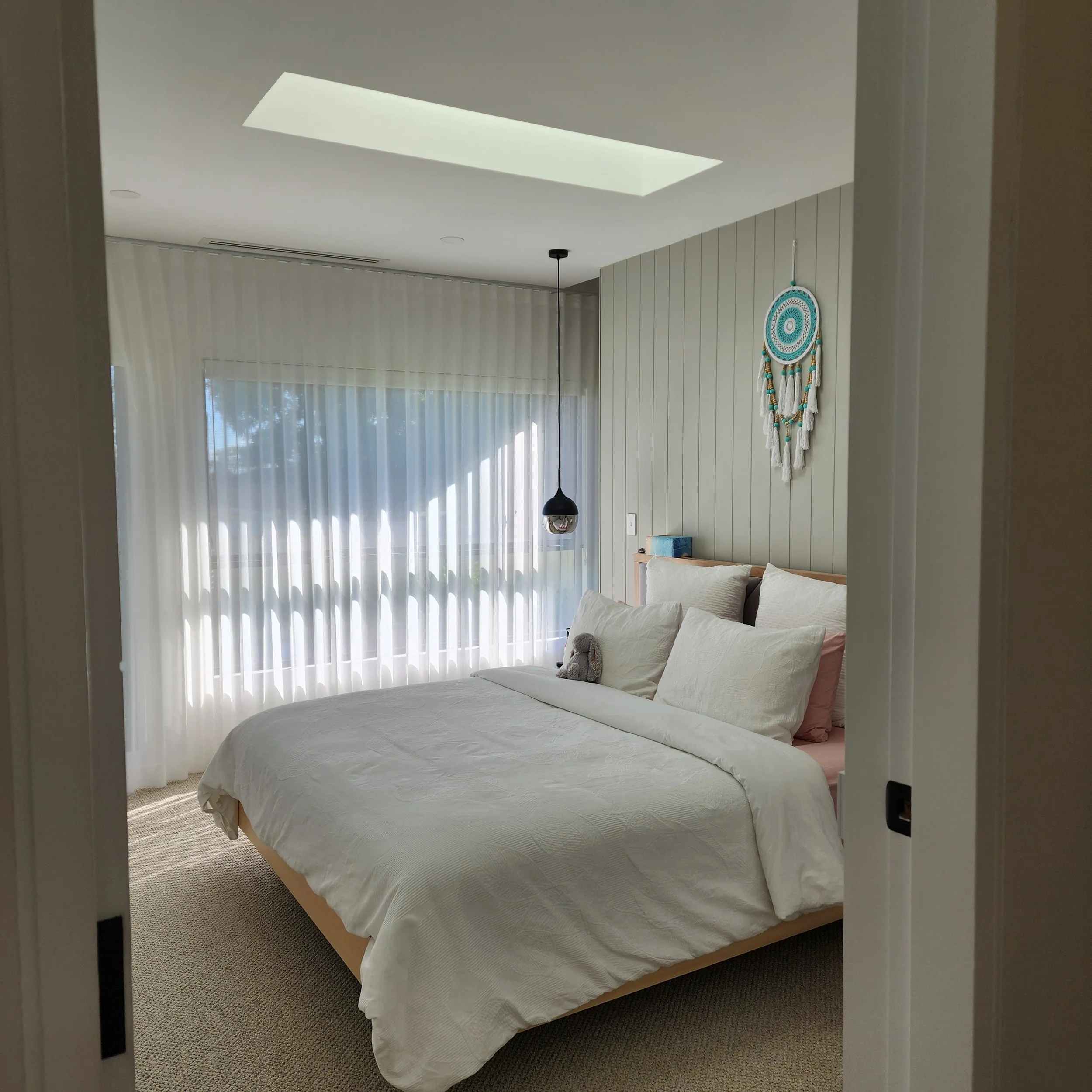 A bedroom with a bed covered in a white quilt, pink pillows, and a plush toy on the bed. The room has large windows with vertical blinds and a hanging light fixture. There's a decorative dreamcatcher on the wall to the right.