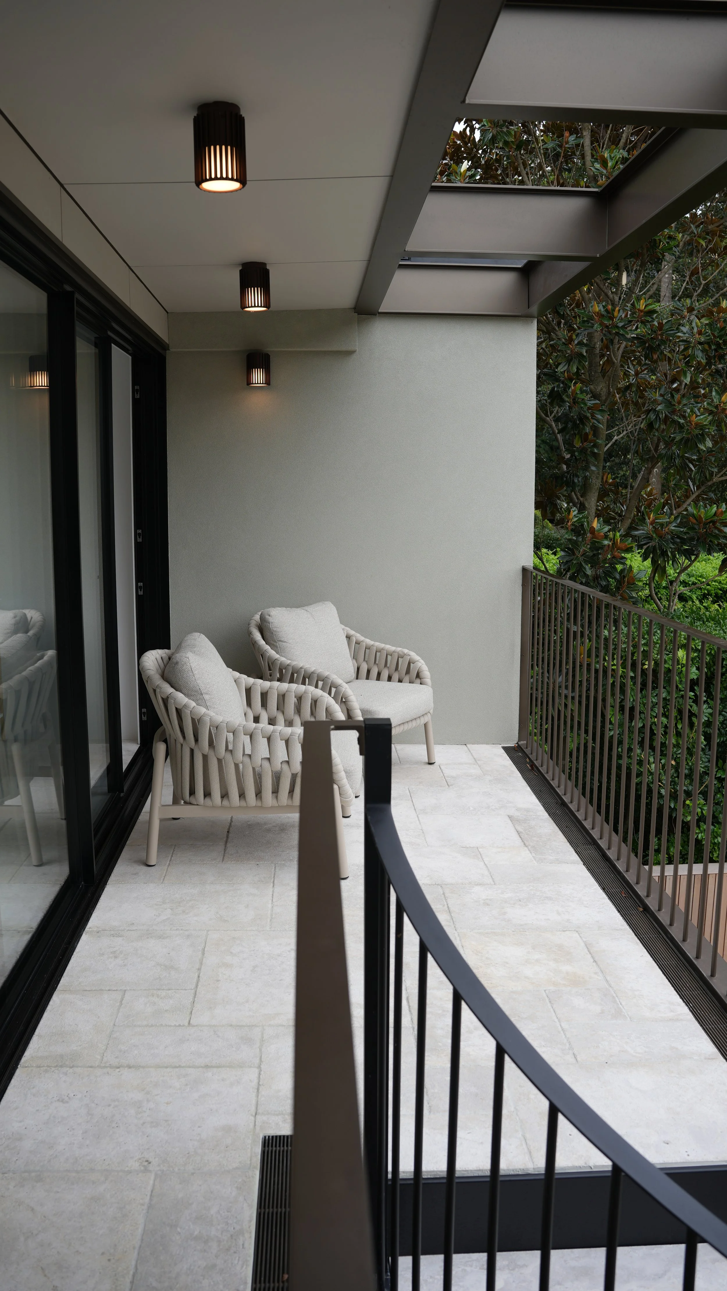 A balcony with two white woven chairs and beige cushions, black metal railing, sliding glass doors, ceiling lights, and view of green trees.