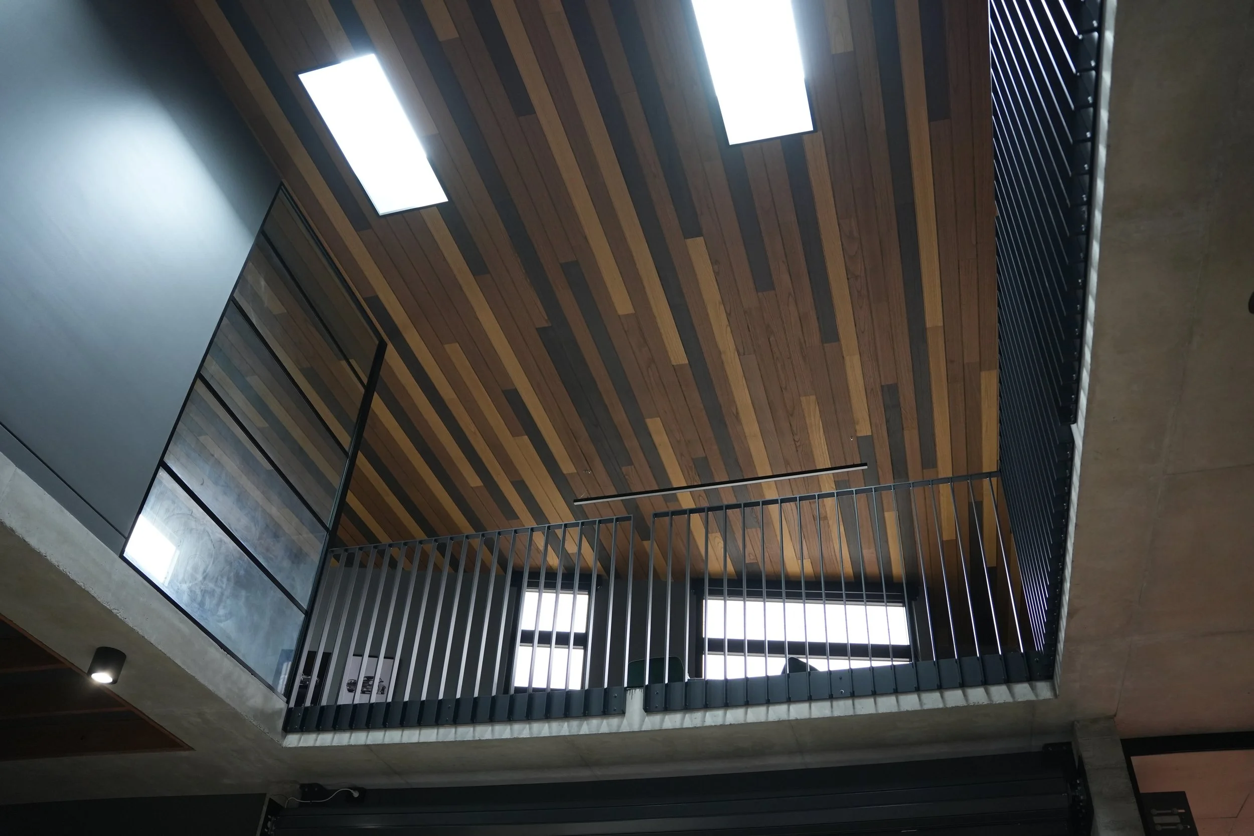 Interior view of a modern building with a wooden ceiling and skylights, viewed from below near a glass railing.