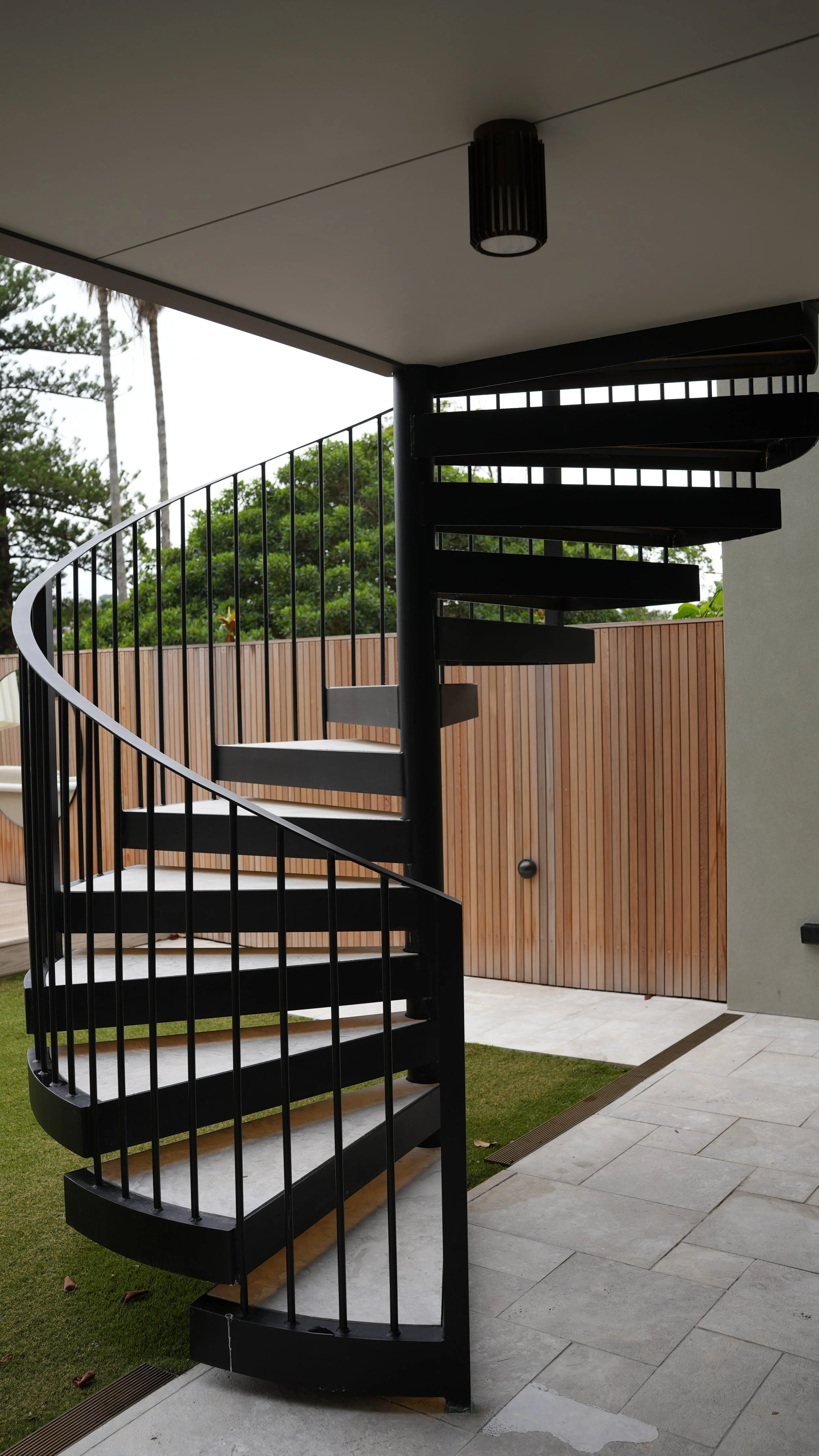 Black spiral staircase next to a wood-paneled fence with trees in the background.