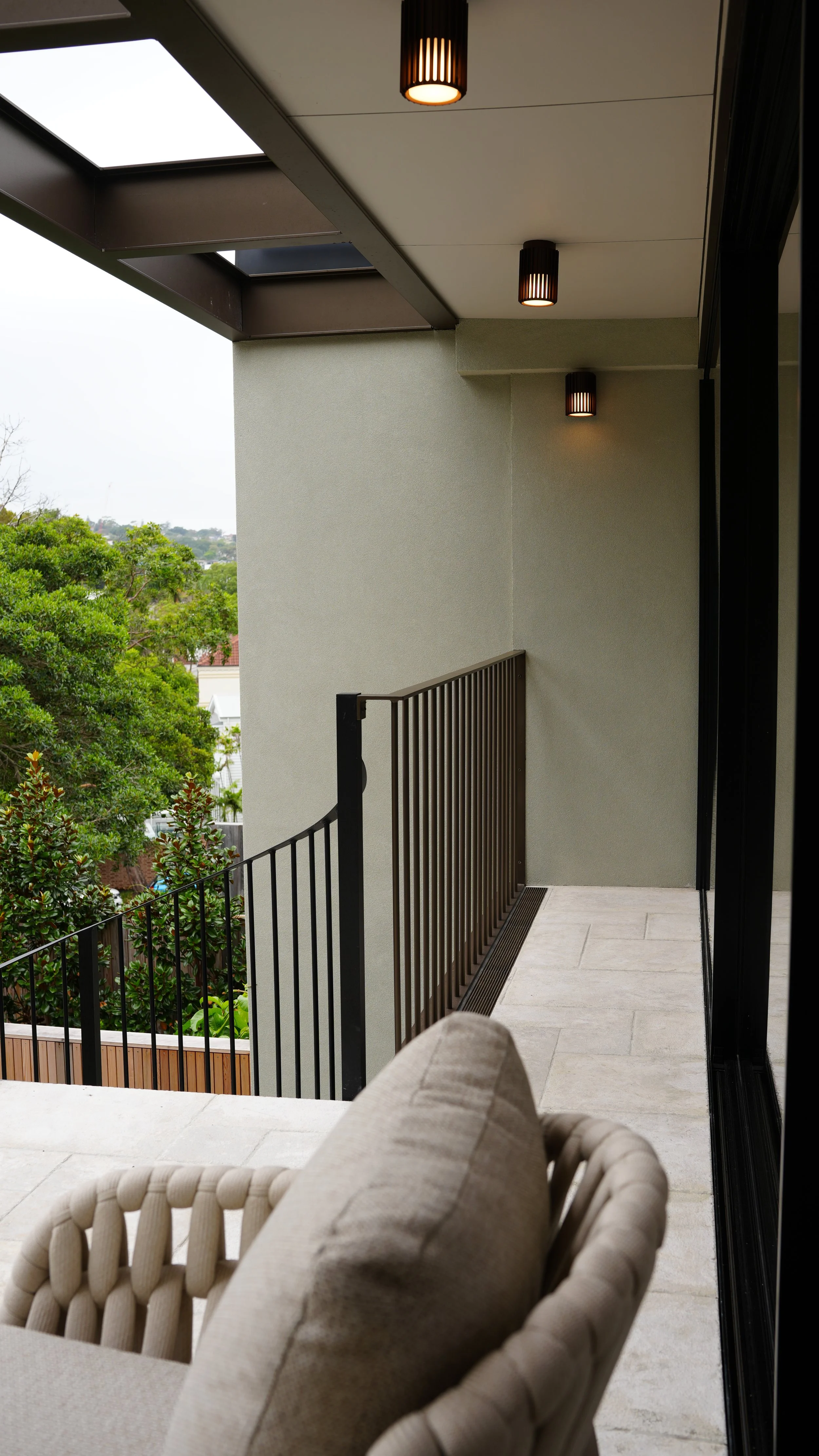 Balcony view with a beige cushioned chair, black metal railing, and outdoor greenery, with a ceiling light and skylight above.