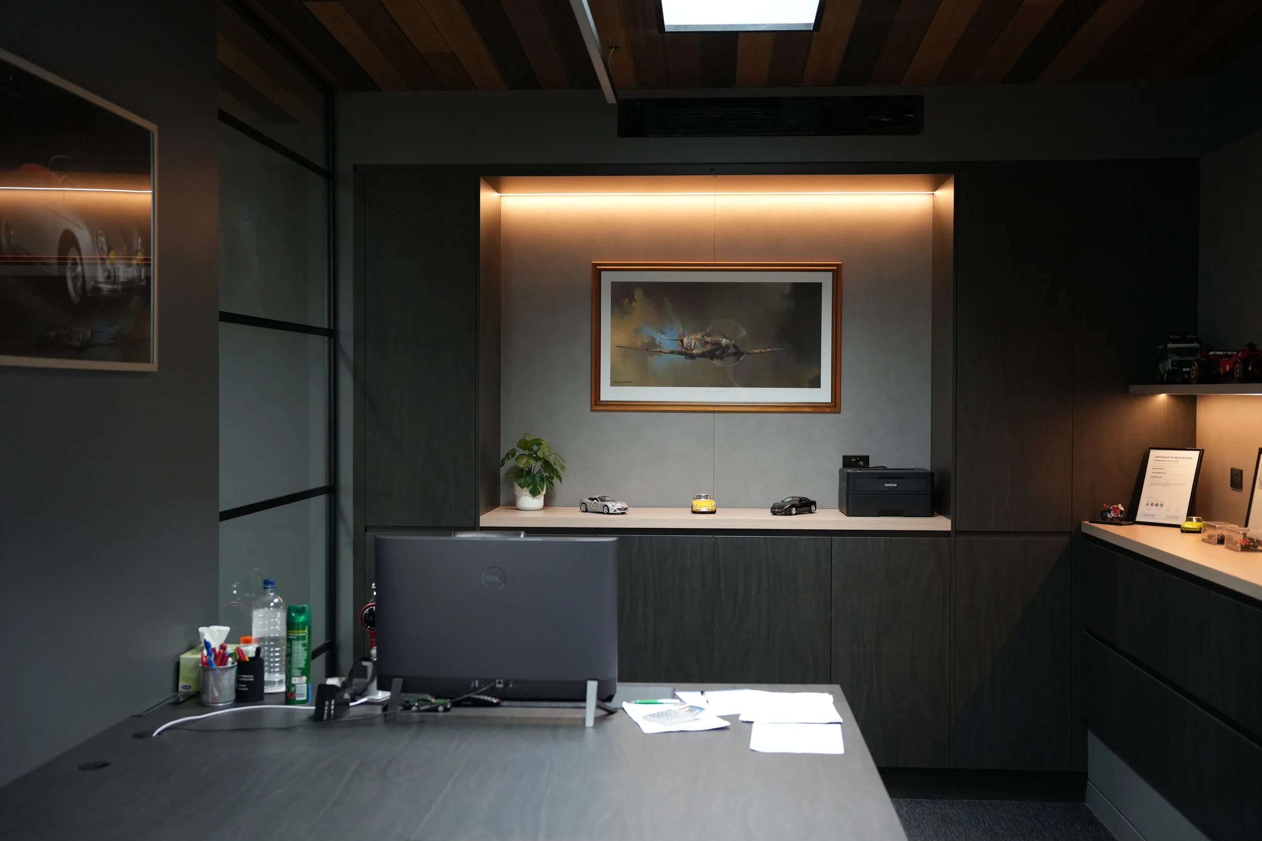 Office desk with scattered papers, pens, and a computer monitor, with a framed airplane painting and model cars on the dark wood cabinets behind.