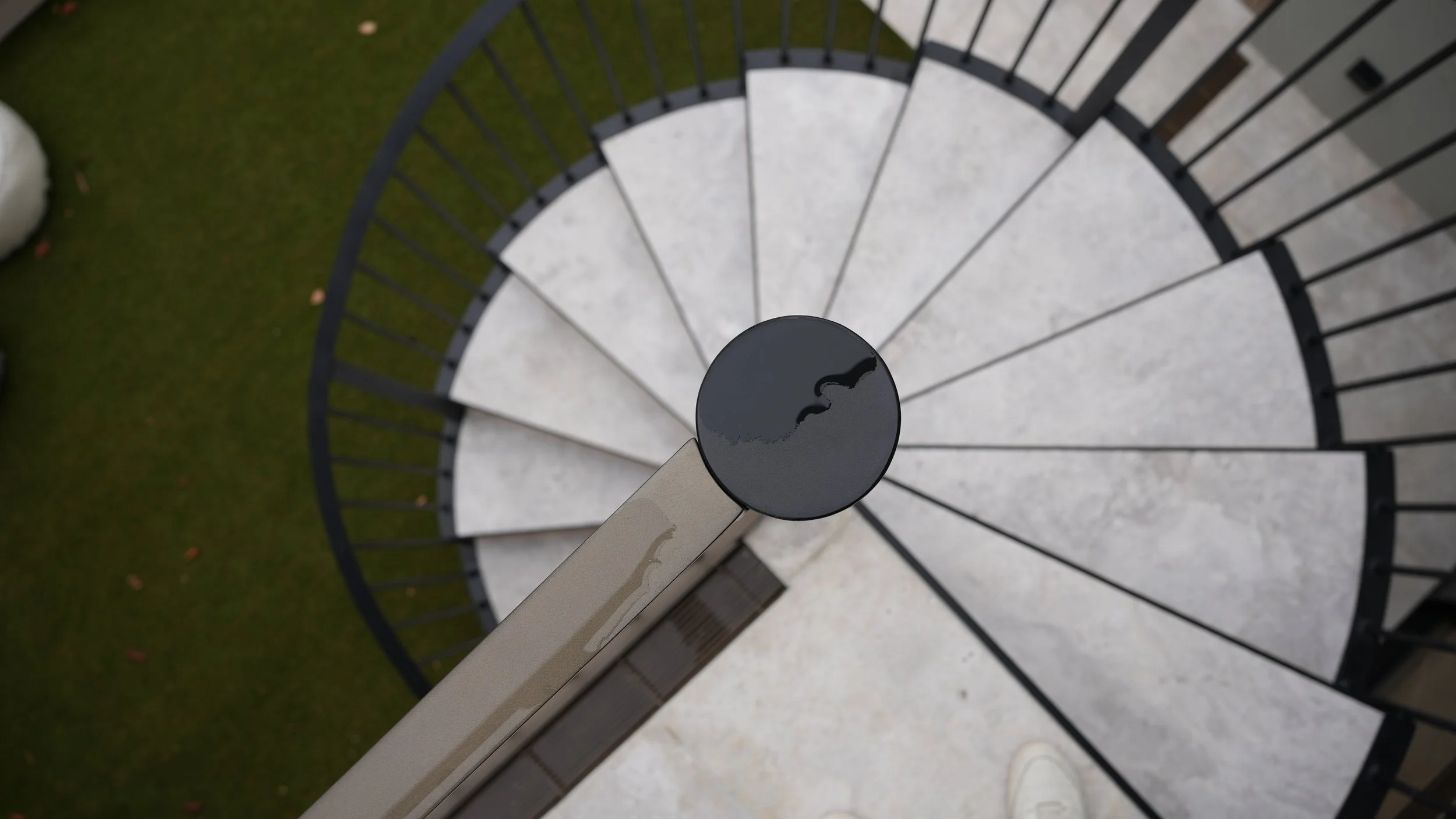 A spiral staircase viewed from above, with a black railing and white steps, outdoors on a green lawn.