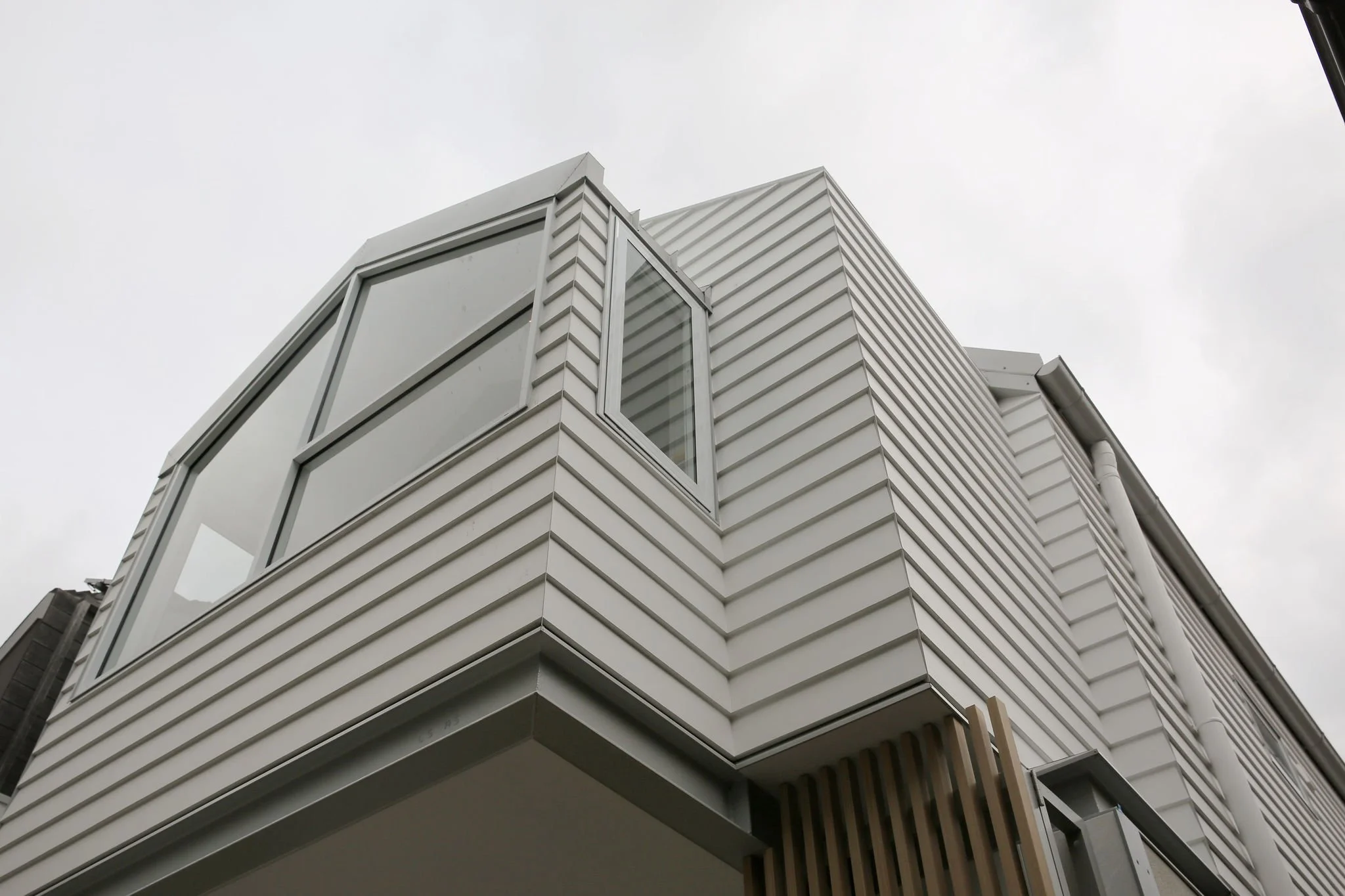 A modern building with white horizontal siding, large glass windows, and a corner with a small balcony. The sky is overcast.