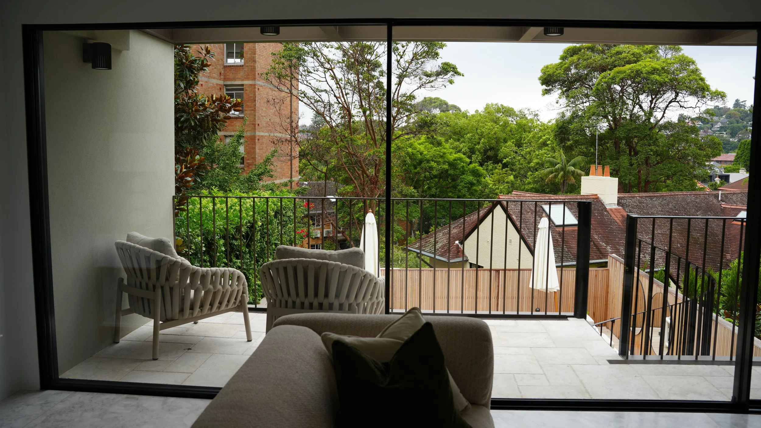 View from indoors of a balcony with white chairs, overlooking green trees and rooftops of neighboring houses with brick and tile roofs.
