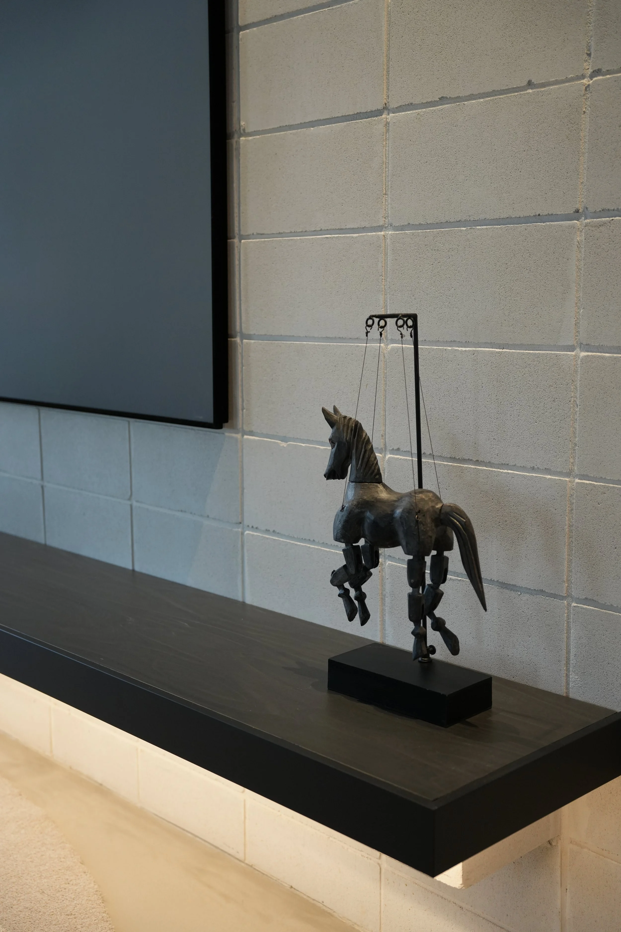 A metal sculpture of a horse on a black rectangular base displayed on a black shelf against a beige brick wall.