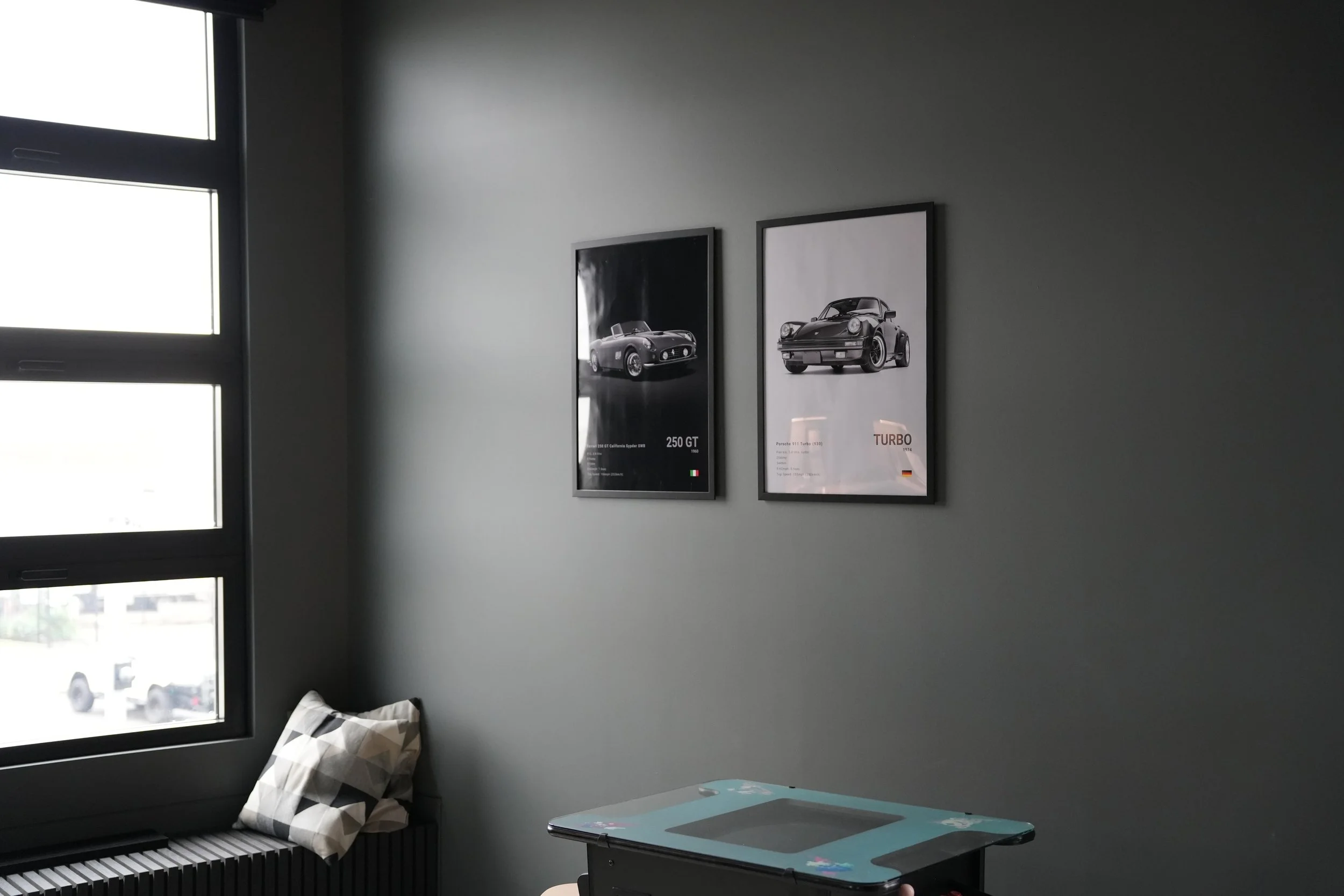 Gray wall with two framed posters of classic Porsche cars, next to a window, with a black and white pillow on a seat below the window, and a blue notebook on a table nearby.