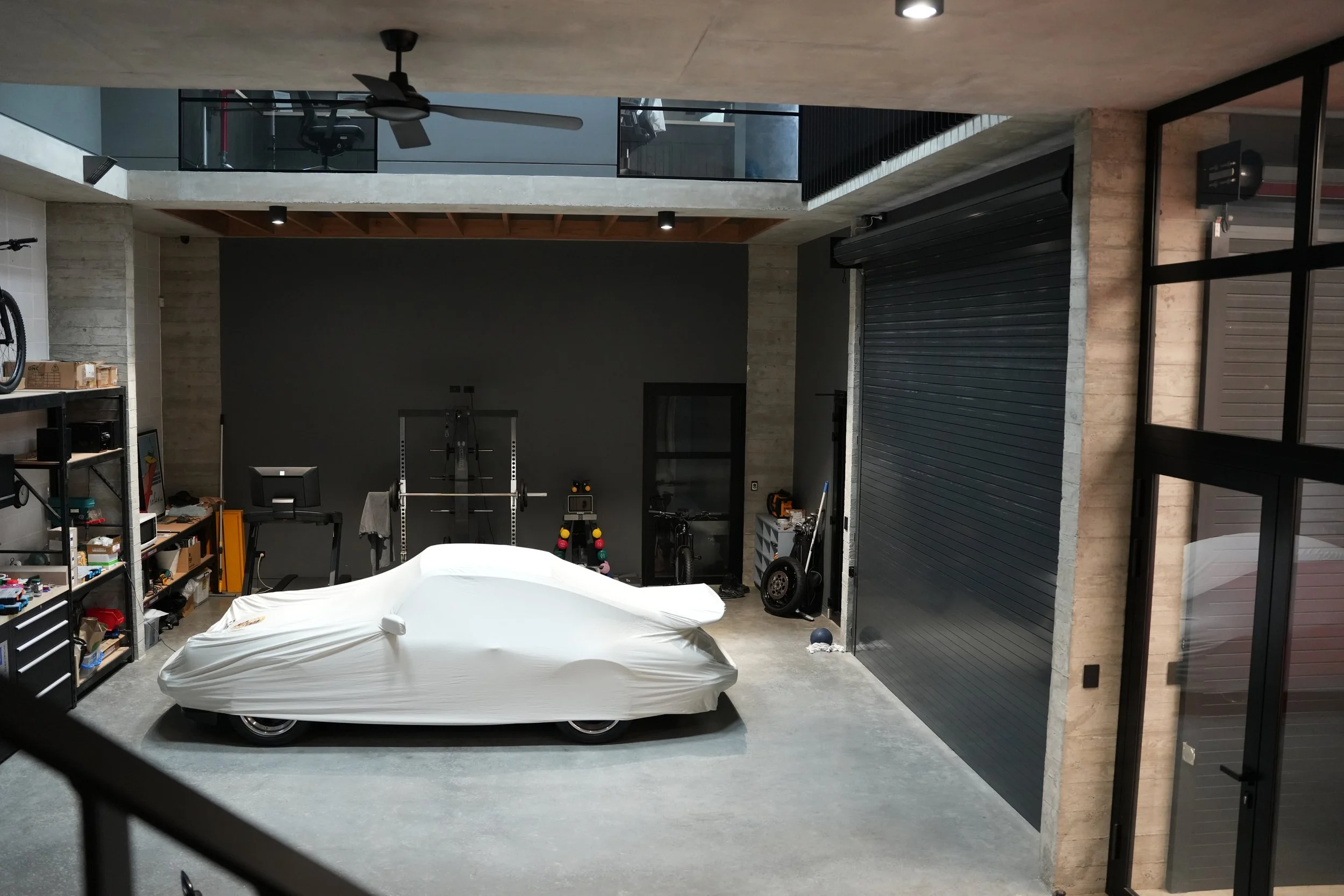 Modern industrial-style garage with a car covered by a white tarp, gym equipment, shelving with boxes and tools, and various items on the concrete floor.