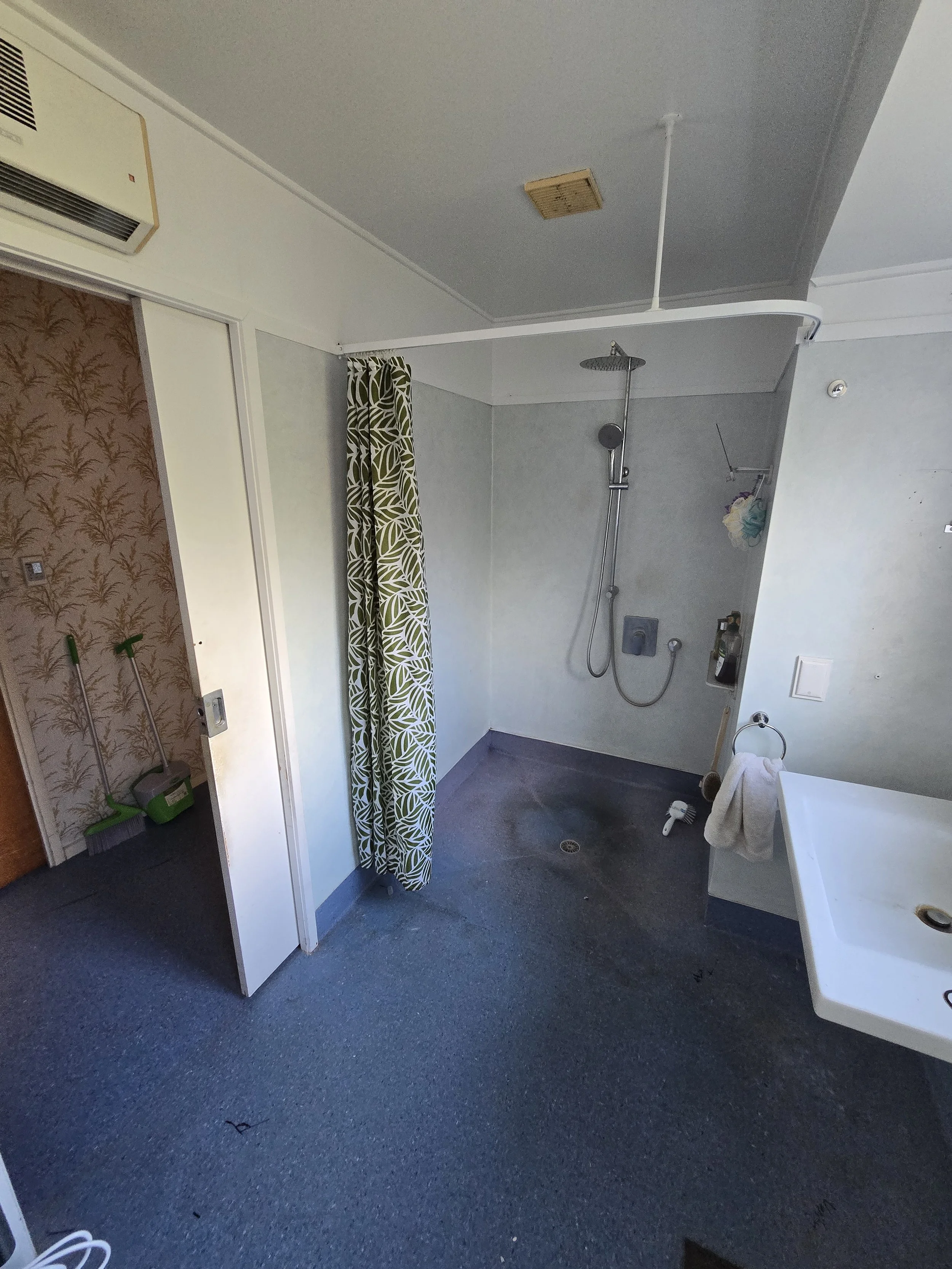 A bathroom with a shower area, a patterned curtain, and cleaning supplies including mops and brooms near the wall.