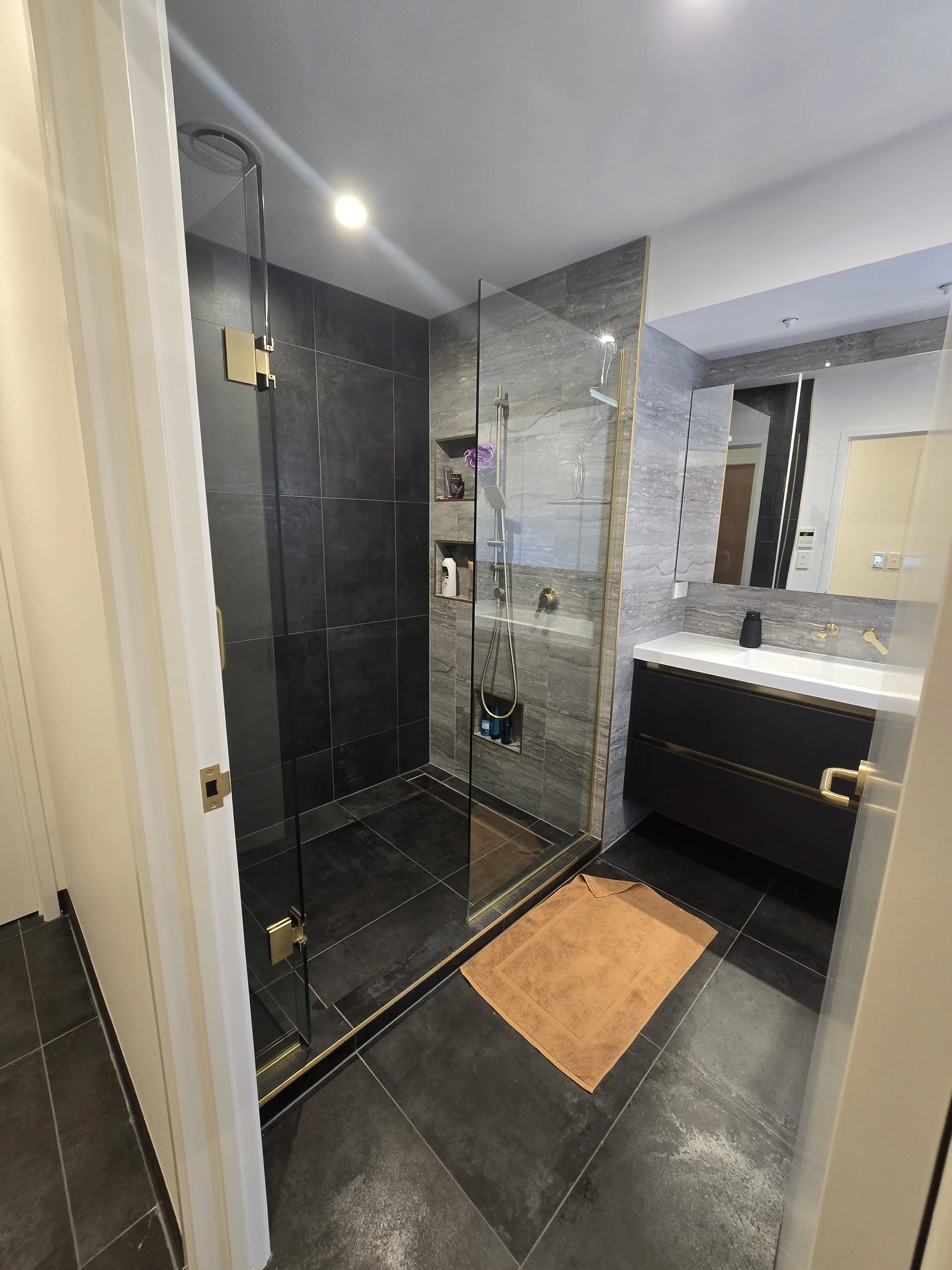 A modern bathroom with a glass-enclosed shower, dark tiled walls and floors, a beige bathmat, and a vanity with a large mirror. The shower area has built-in shelves with toiletries and an orchid flower.