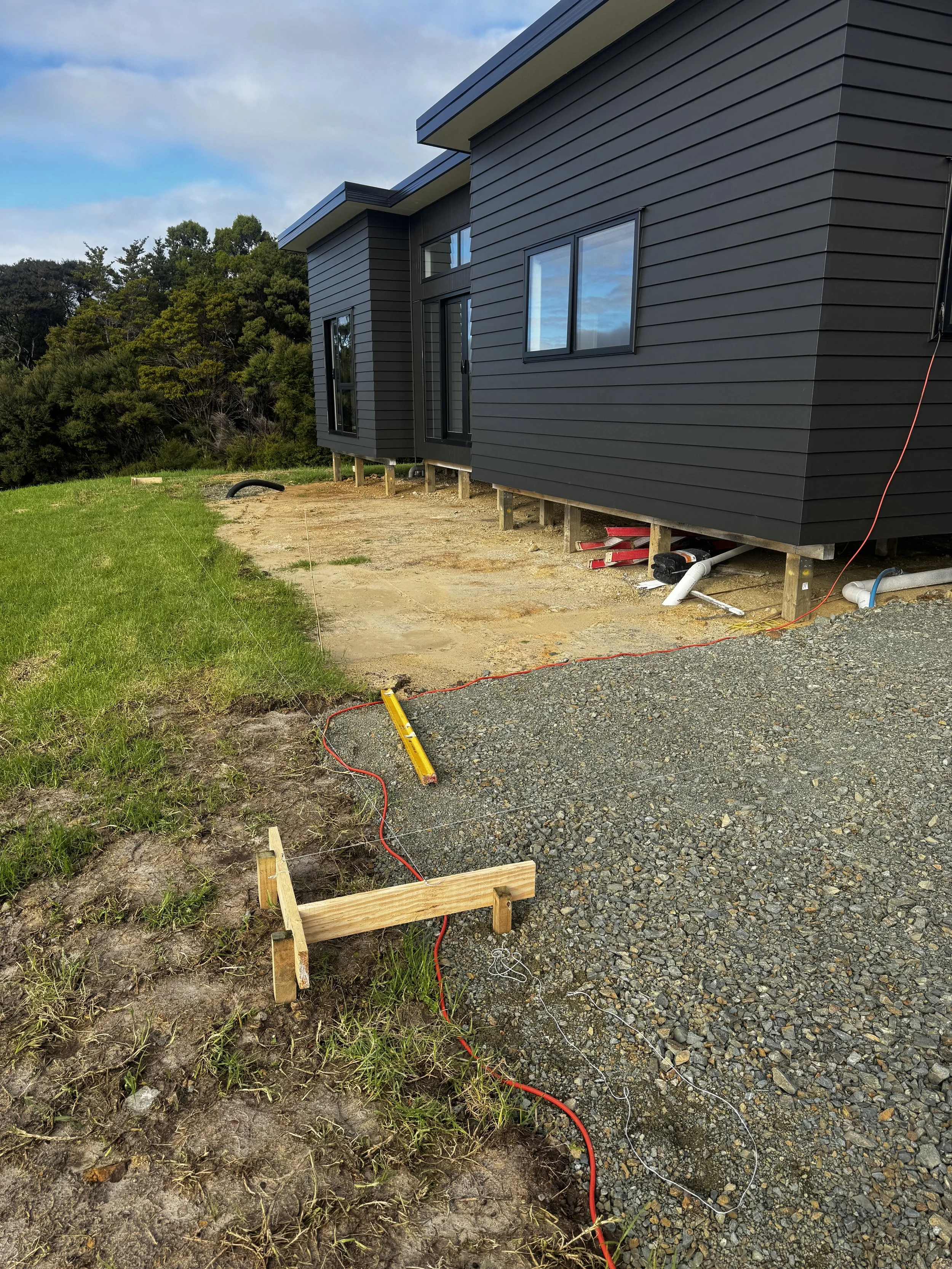 Construction site with a dark gray modern house on stilts, electrical wiring, and a gravel path under construction, with a grassy area and trees in the background.