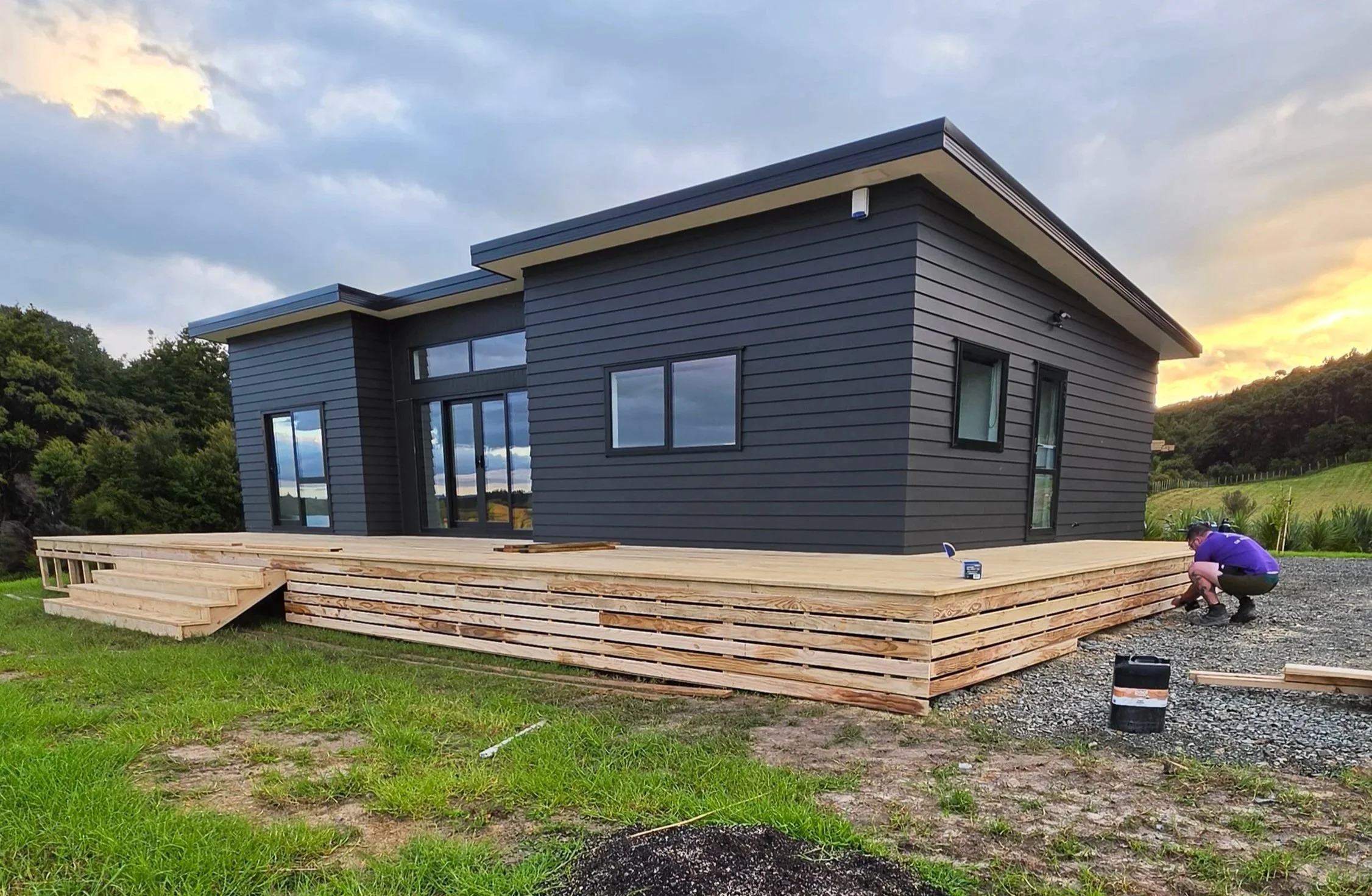 A modern house with dark gray siding and large windows is under construction on a green lawn. The house has a sloped roof, and a person is kneeling on the wooden deck, which is in the process of being built, working on the foundation or framing. The background features a cloudy sky and distant trees.