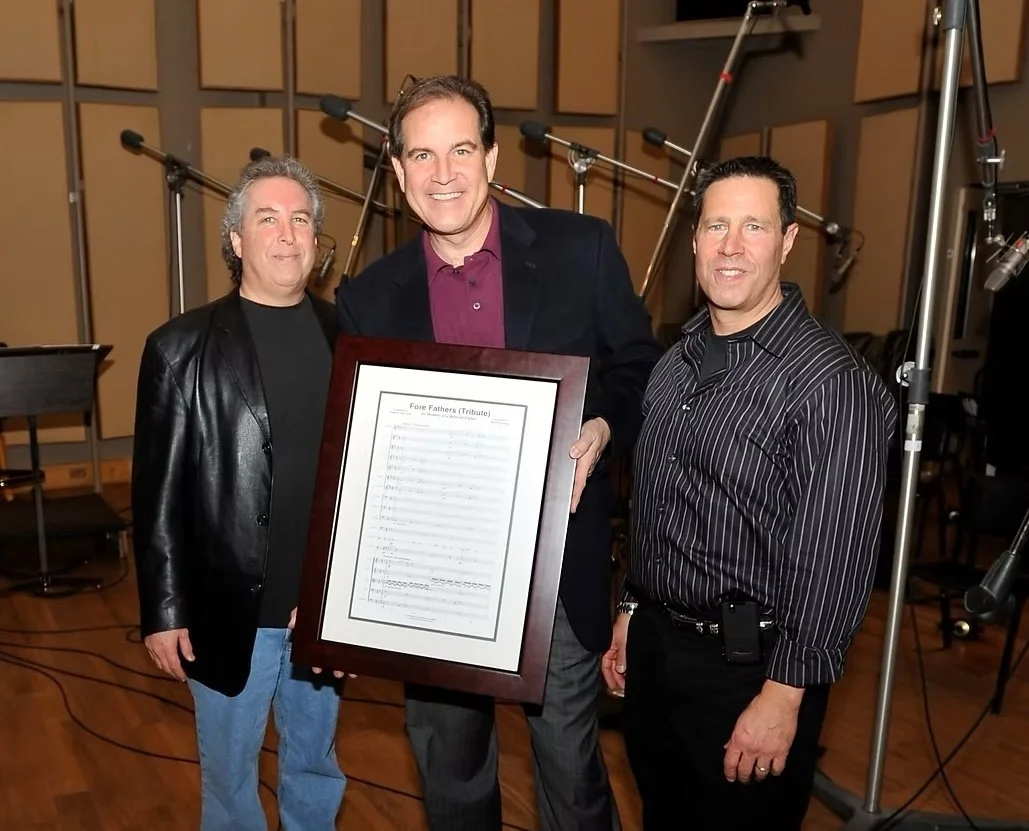 Three men standing together in a recording studio, with one holding a framed sheet of music titled "Fore Fathers (Tribute)."