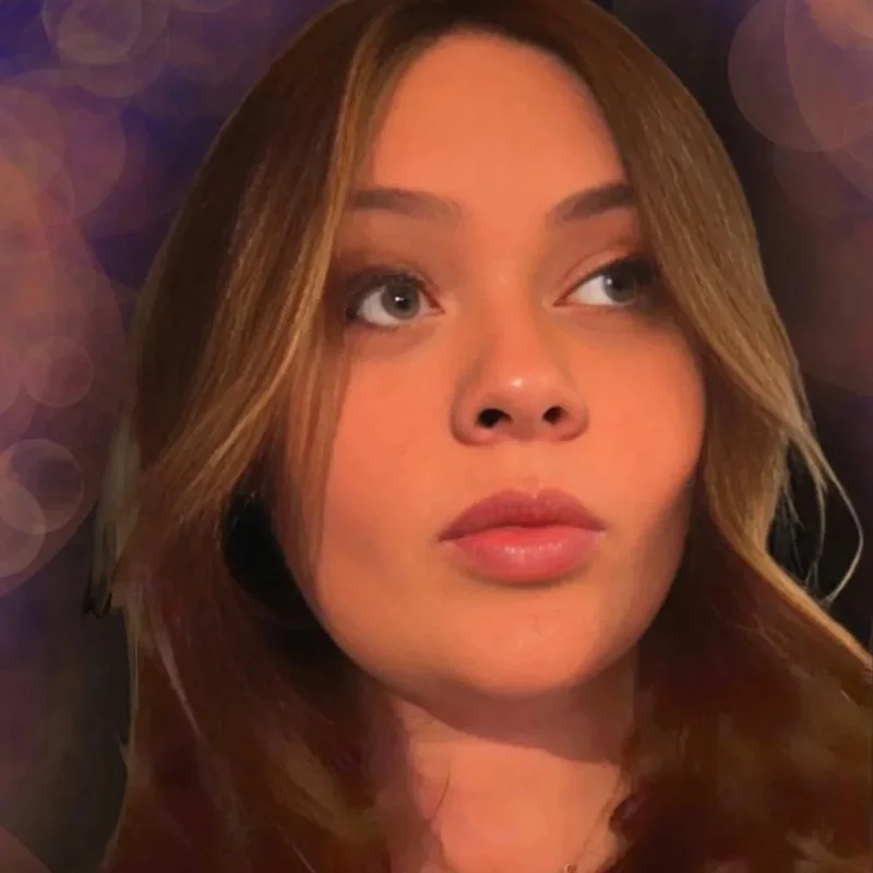 A young woman with light brown hair, blue eyes, and makeup, looking to the side with a neutral expression. The background has soft, circular bokeh lights.