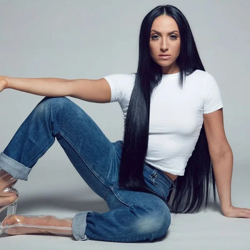 A woman with long black hair wearing a white t-shirt and blue jeans, sitting on the floor with one arm extended and the other supporting her weight, against a plain gray background.