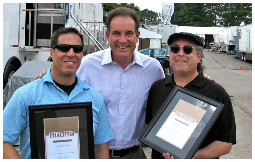 Three men smiling outdoors, two holding framed certificates, with trailers and equipment in the background.