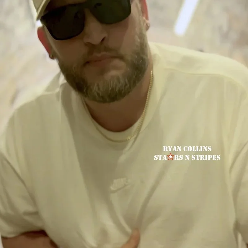 Close-up of a man wearing black sunglasses, a beige cap, a gold chain, and a cream-colored t-shirt with the text 'Ryan Collins Stars N Stripes'.