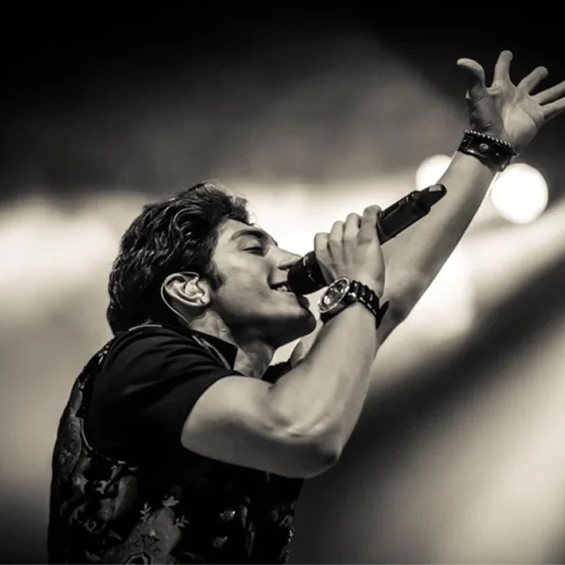 A male singer passionately performing on stage, holding a microphone close to his mouth with his eyes closed and one arm raised in the air.